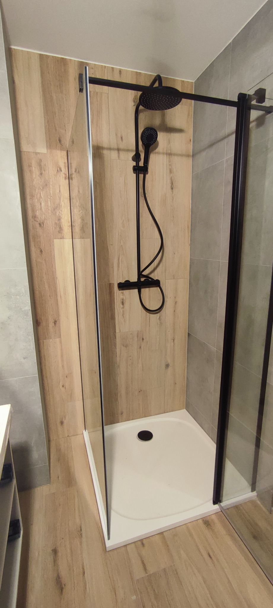 Nowoczesna kabina prysznicowa z czarnymi profilami i deszczownicą, wykończona płytkami imitującymi drewno i szare płytki.