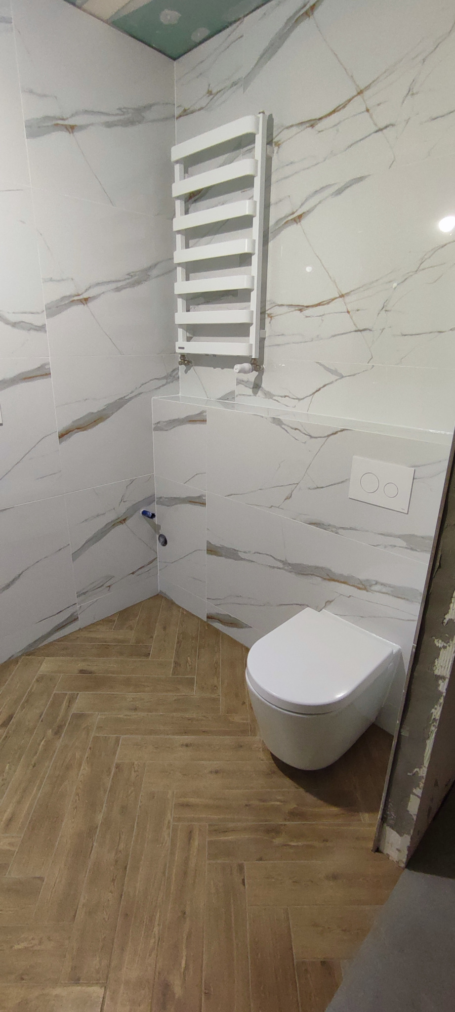 Nowoczesna toaleta wisząca w łazience wykończonej białymi płytkami imitującymi marmur z brązowymi żyłami, drewnopodobna podłoga ułożona w jodełkę, biały grzejnik drabinkowy.