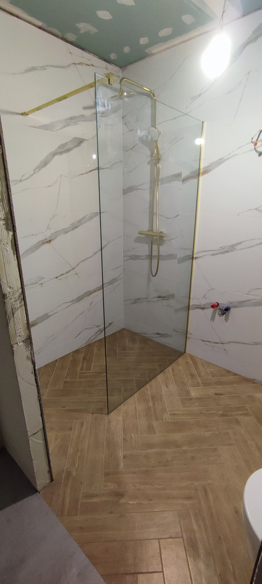 Nowoczesna kabina prysznicowa walk-in z złoconymi elementami i marmurowymi płytkami w łazience w Zielonej Górze, widok na ułożoną w jodełkę podłogę z drewnopodobnych płytek.