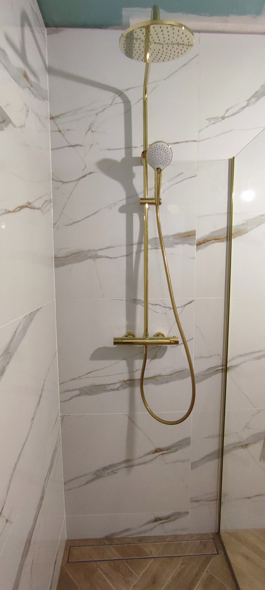 Złota bateria prysznicowa z deszczownicą i słuchawką na tle marmurowych płytek w kabinie prysznicowej z odpływem liniowym w podłodze ułożonej w jodełkę.