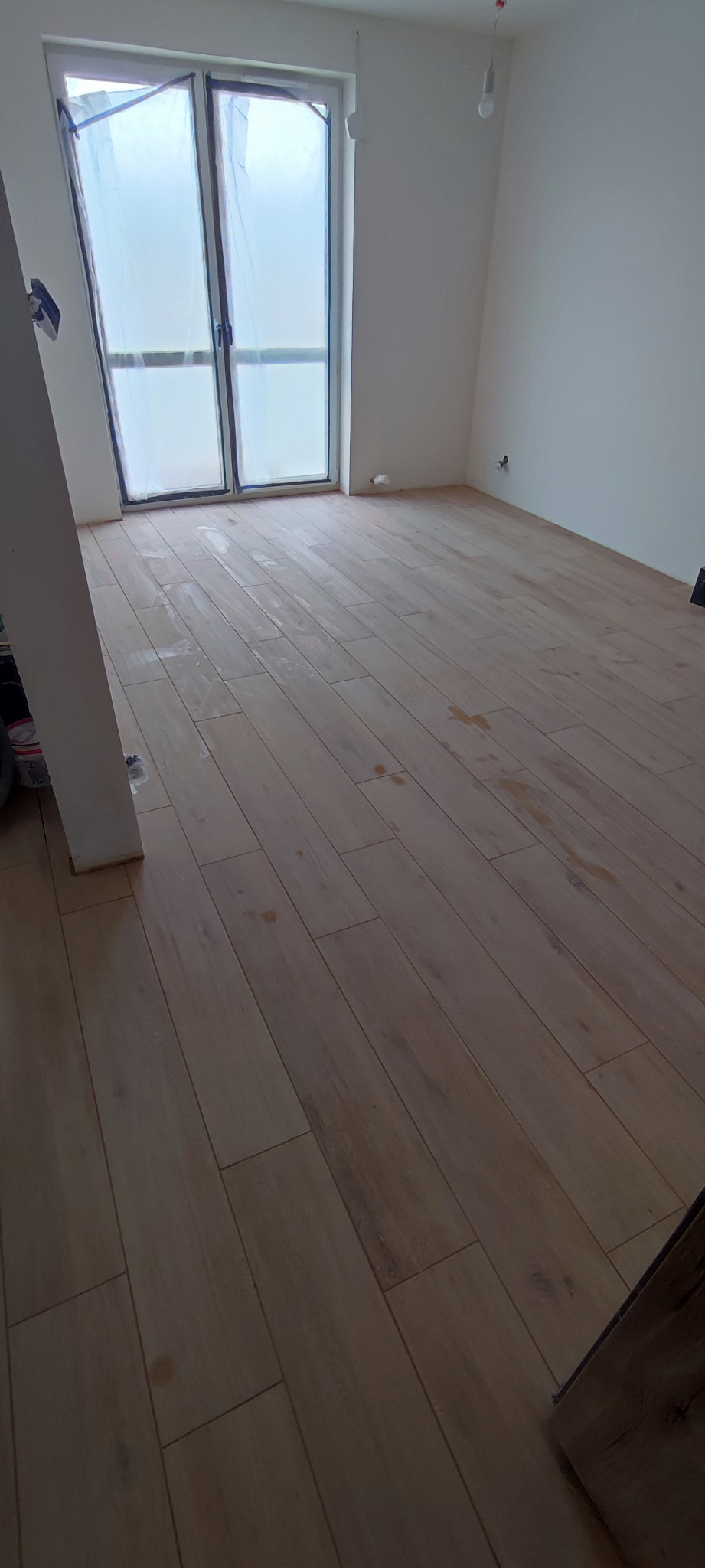 Jasne panele podłogowe ułożone w nowym pokoju, okno zabezpieczone folią malarską, widoczne drobne zabrudzenia na powierzchni podłogi.