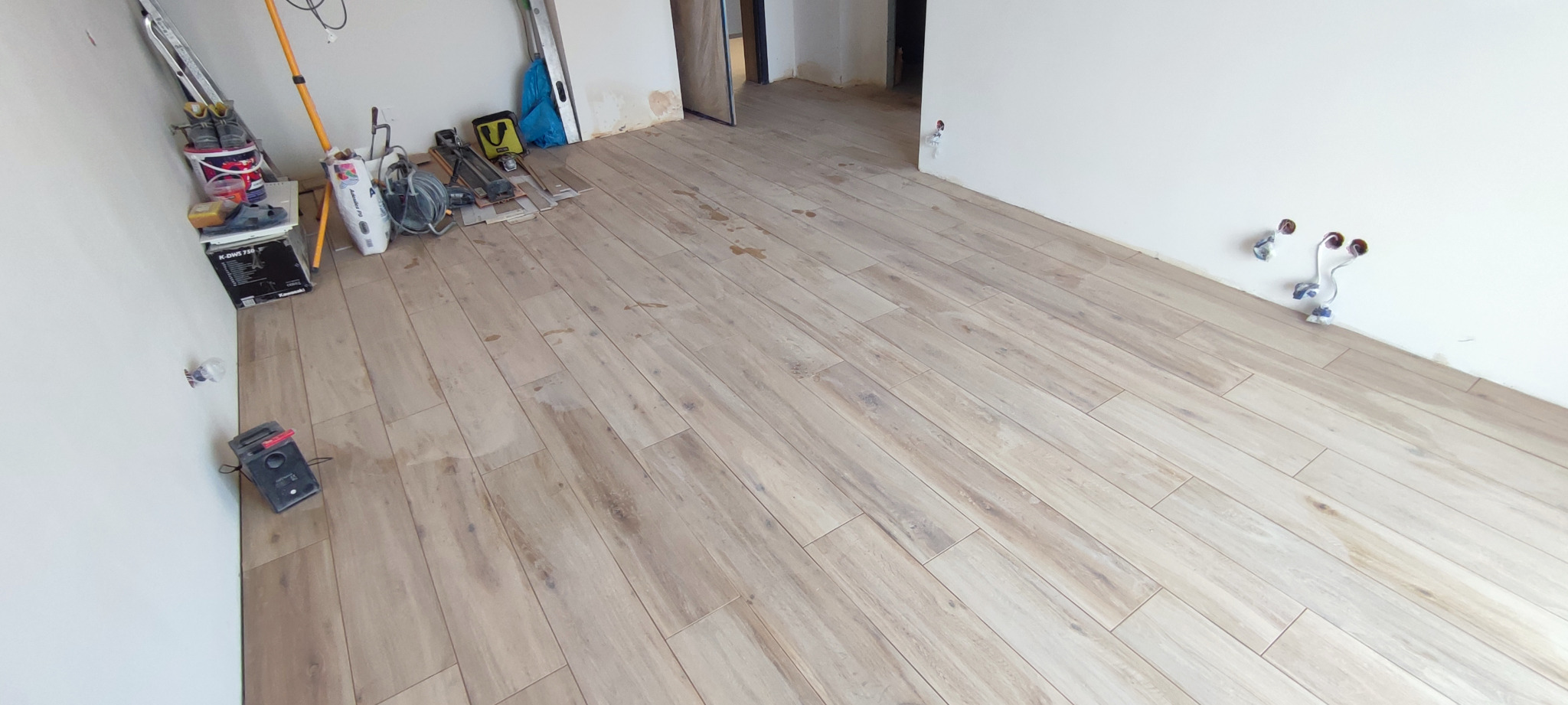 Ułożona podłoga z paneli drewnopodobnych w jasnym odcieniu, widoczne narzędzia glazurnicze i niezabudowane instalacje na ścianie.