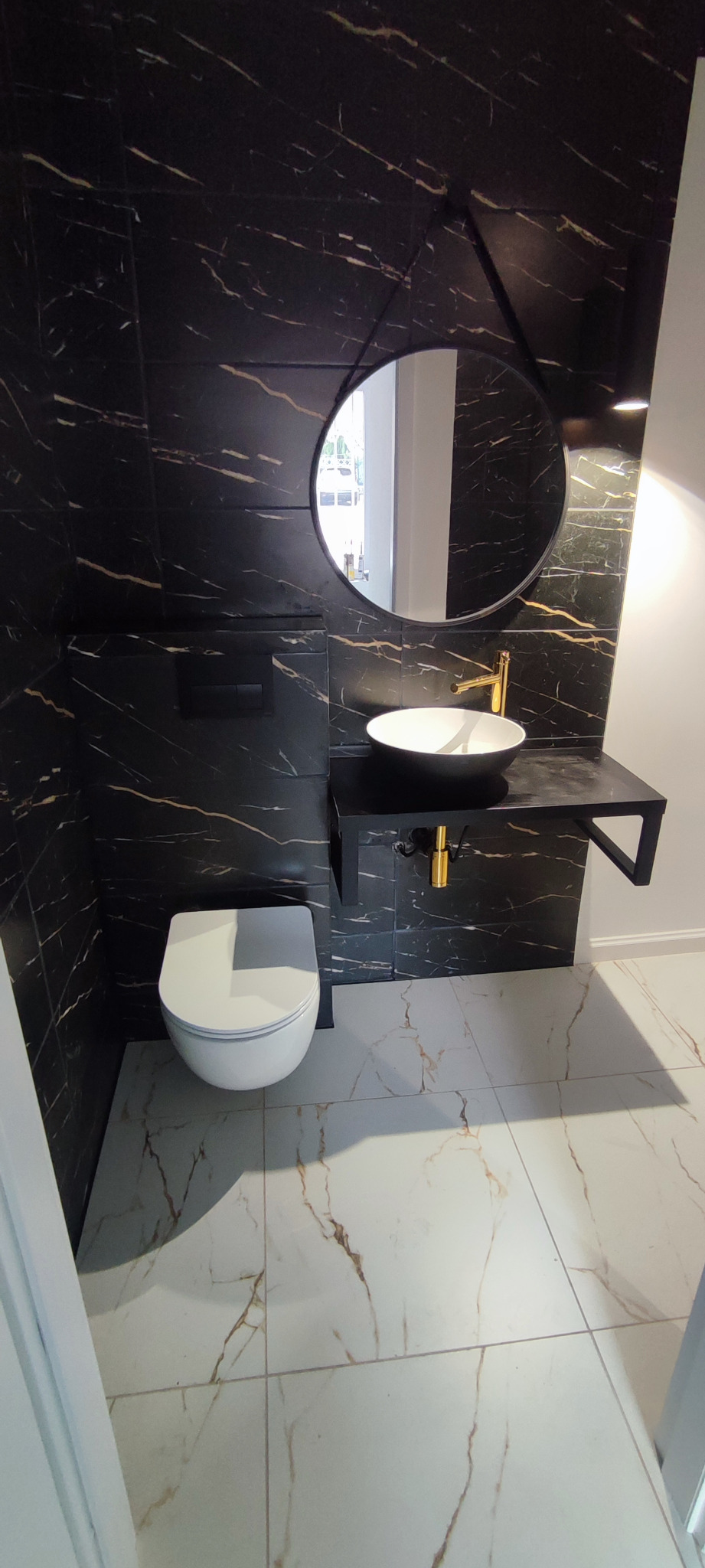 Nowoczesna łazienka z czarnymi płytkami imitującymi marmur ze złotymi żyłami na ścianach, okrągłe lustro, biała umywalka nablatowa z złotą armaturą i podwieszana miska WC.