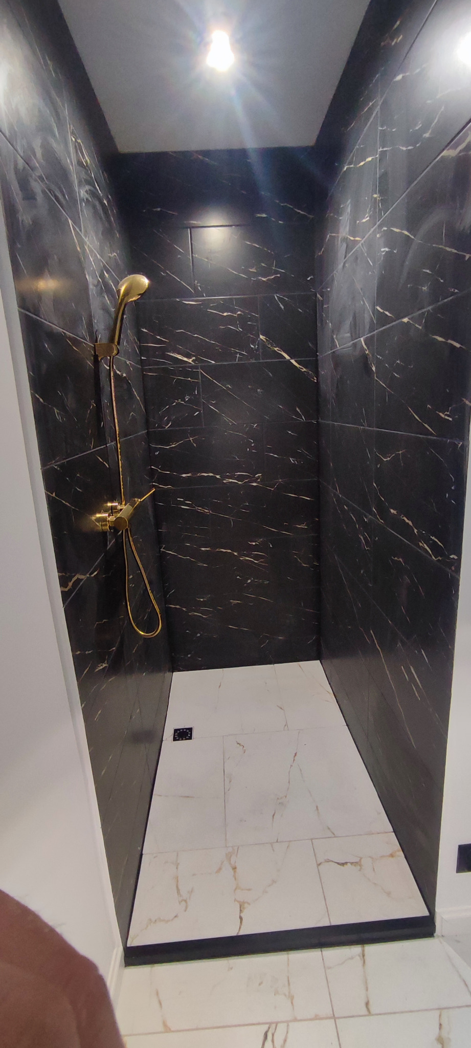 Kabina prysznicowa wykończona czarnymi płytkami imitującymi marmur ze złotymi żyłami, złota armatura, biała podłoga z marmurowym wzorem, czarny odpływ liniowy.