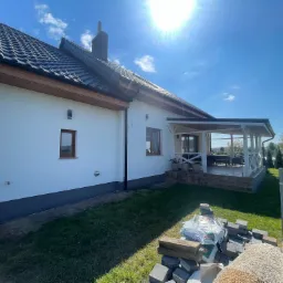 Biały dom jednorodzinny z ciemną dachówką i drewnianymi elementami wykończeniowymi, widok z boku, z przygotowanym materiałem budowlanym do wykończenia ogrodu, kostka brukowa i worki z ziemią...