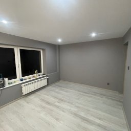 Zakład ogólnobudowlany Damian Pawlak - Pokój po odświeżeniu: szare ściany, jasna podłoga imitująca drewno, biały grzejnik pod oknem, narzędzia na parapecie.