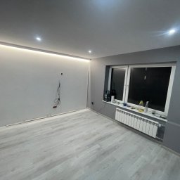 Zakład ogólnobudowlany Damian Pawlak - Szare, odnowione wnętrze pokoju z drewnopodobną podłogą, białym grzejnikiem pod oknem i podświetleniem LED pod sufitem, widoczne wystające kable elektryczne.