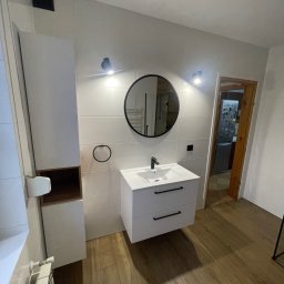 Zakład ogólnobudowlany Damian Pawlak - Nowoczesna łazienka w Śremie po liftingu, z białymi płytkami, drewnopodobną podłogą, okrągłym lustrem i minimalistyczną szafką z umywalką.