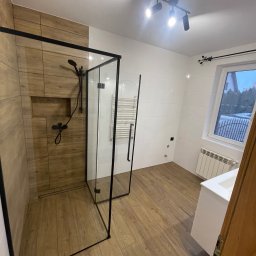 Zakład ogólnobudowlany Damian Pawlak - Nowoczesna łazienka z kabiną prysznicową typu walk-in z czarnymi profilami, drewnopodobne płytki w strefie prysznica i na podłodze, białe płytki na pozostałych ścianach, grzejnik drabinkowy, biała...