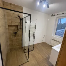 Nowoczesna łazienka z kabiną prysznicową typu walk-in z czarnymi profilami, drewnopodobne płytki w strefie prysznica i na podłodze, białe płytki na pozostałych ścianach, grzejnik drabinkowy, biała...