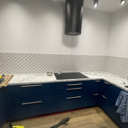 Zakład ogólnobudowlany Damian Pawlak - Niebieskie szafki kuchenne z marmurowym blatem i płytkami mozaikowymi na ścianie, czarny okap, narzędzia glazurnicze na blacie, widoczne etapy prac wykończeniowych.