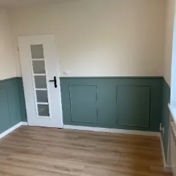 Wnętrze pokoju po remoncie: zielona lamperia na ścianach, drewnopodobna podłoga, białe drzwi z szybami z motywem dmuchawca.