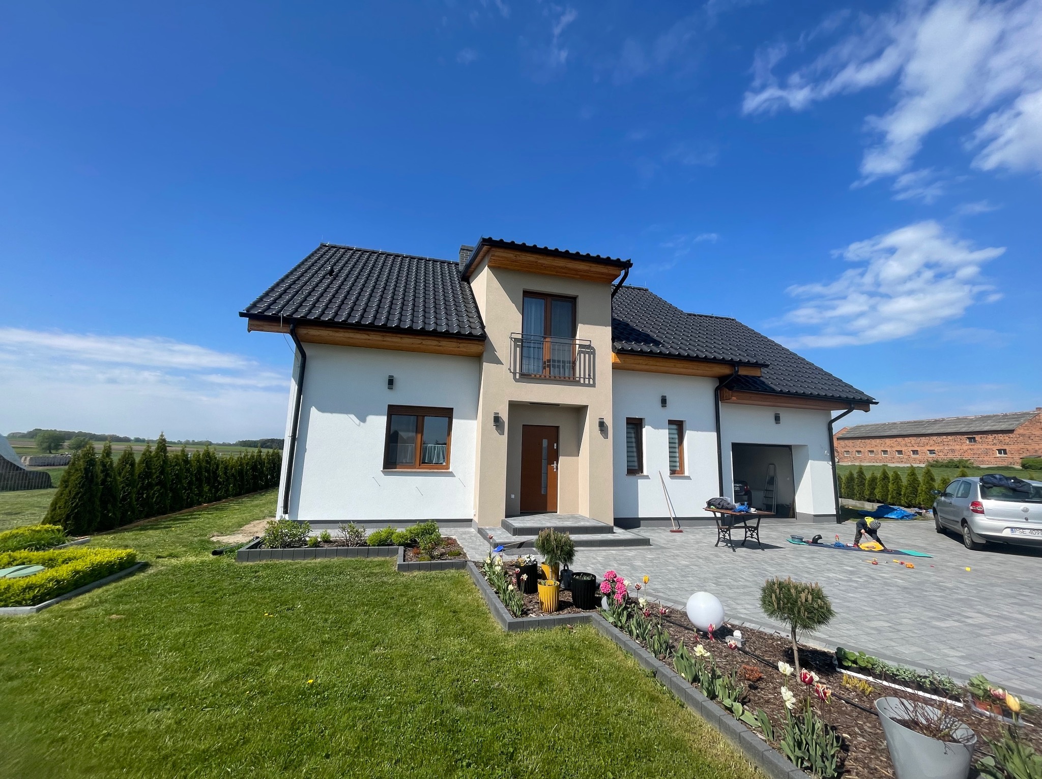 Nowoczesny dom jednorodzinny z dwuspadowym dachem i balkonem, otoczony zadbanym ogrodem z kostką brukową i rabatami kwiatowymi, widok z przodu w słoneczny dzień.