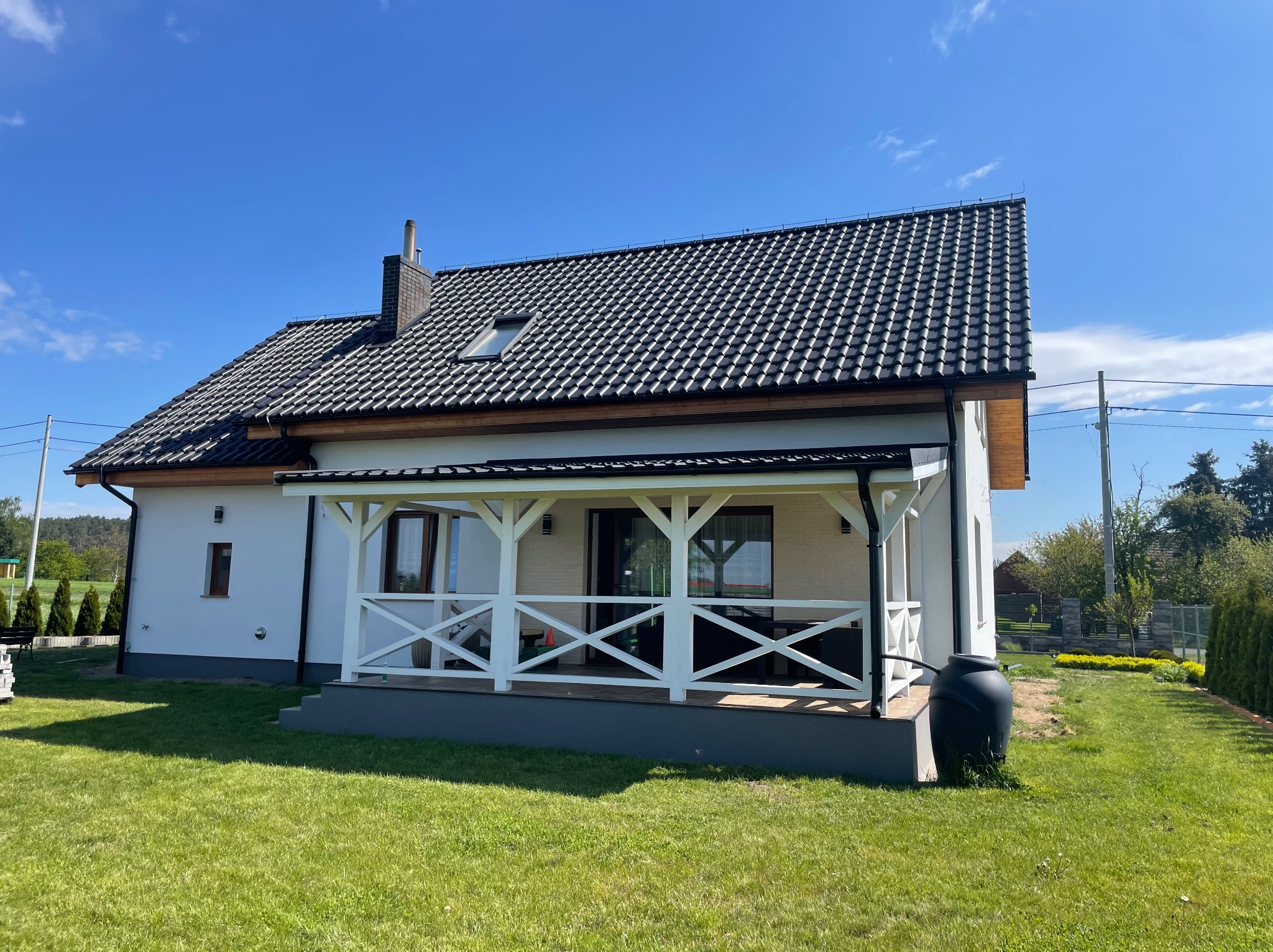 Biały dom jednorodzinny z ciemną dachówką i drewnianą werandą z białymi barierkami w kształcie krzyży, widok z ogrodu w słoneczny dzień.