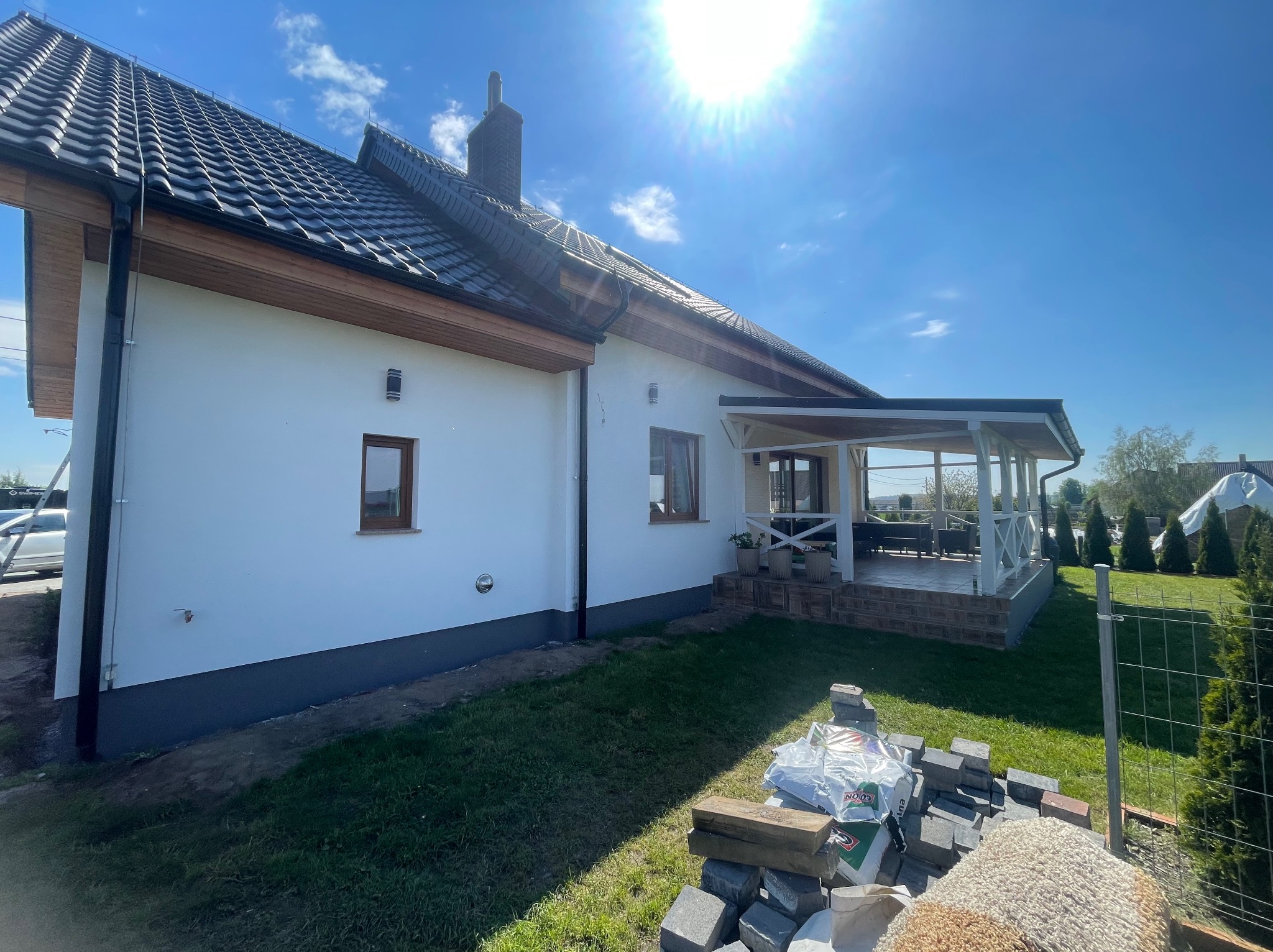 Biały dom jednorodzinny z ciemną dachówką i drewnianymi elementami wykończeniowymi, widok z boku, z przygotowanym materiałem budowlanym do wykończenia ogrodu, kostka brukowa i worki z ziemią...