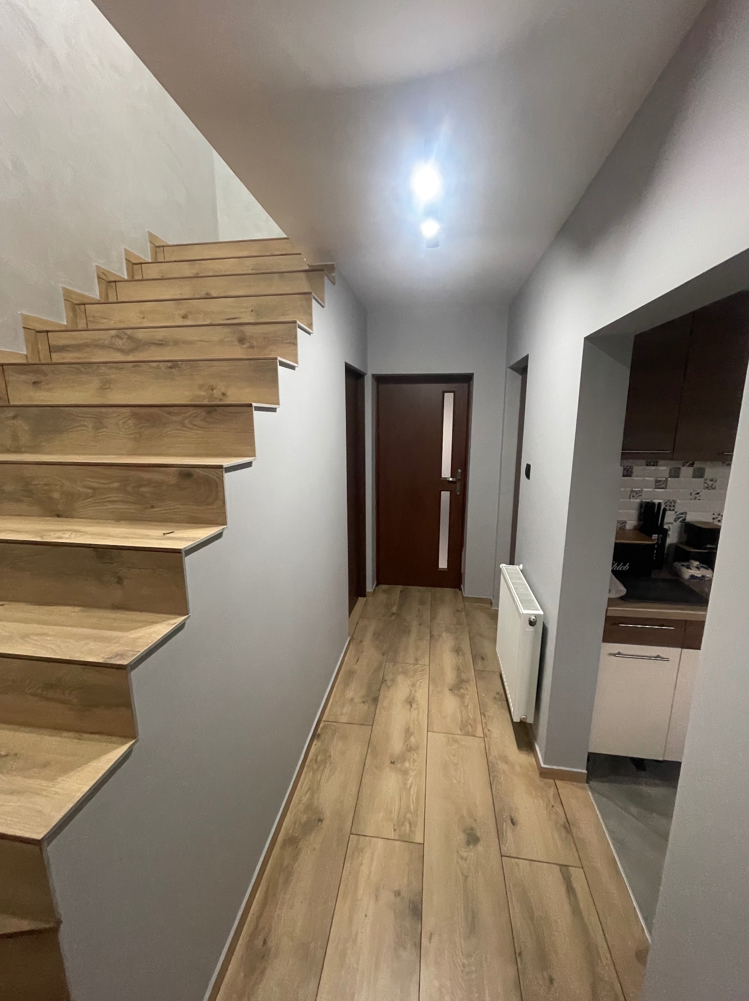Korytarz z drewnopodobnymi płytkami na podłodze i schodach, szare ściany, biały grzejnik oraz widok na kuchnię z szaro-białymi szafkami i brązowymi szafkami wiszącymi.