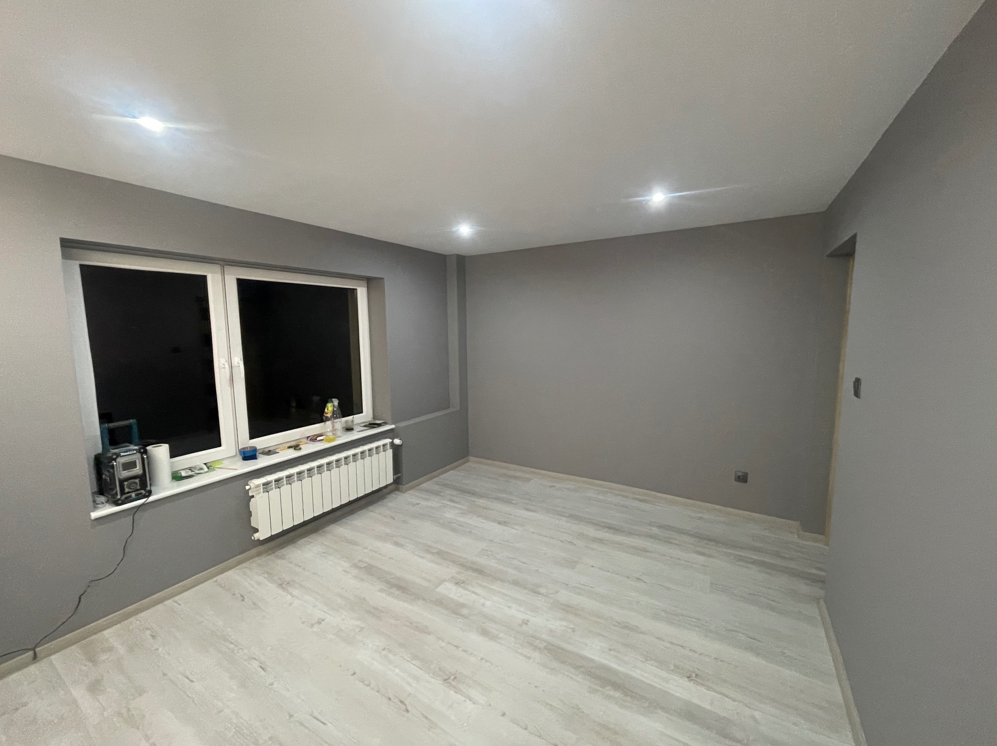 Pokój po odświeżeniu: szare ściany, jasna podłoga imitująca drewno, biały grzejnik pod oknem, narzędzia na parapecie.