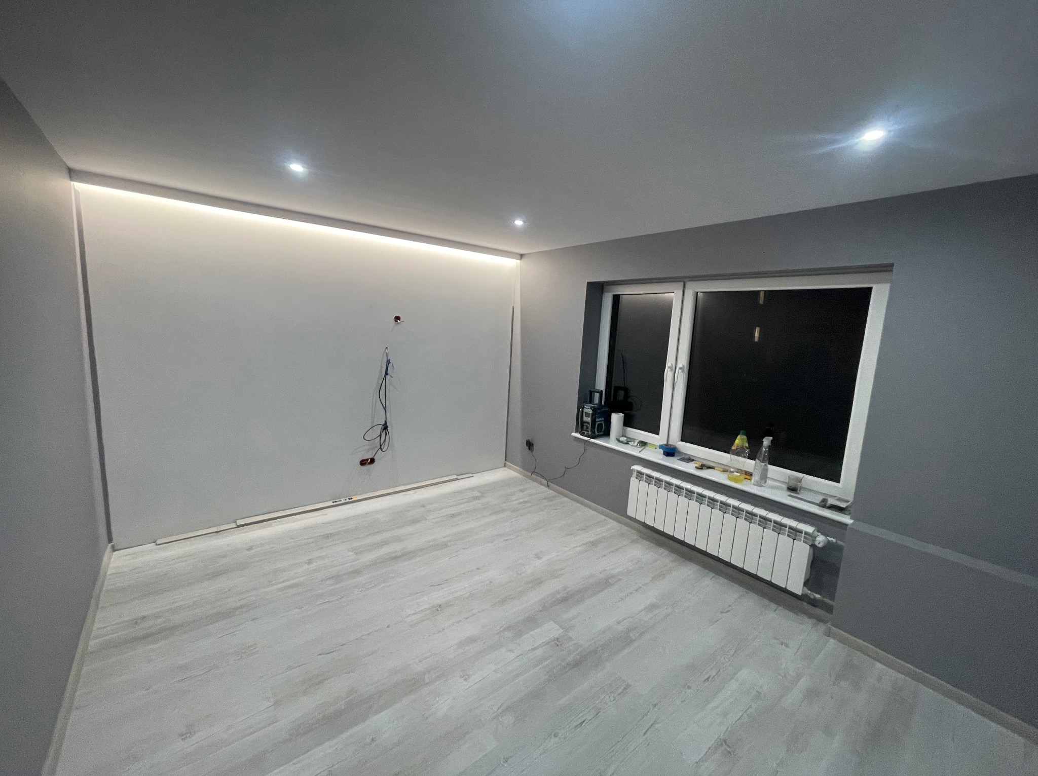 Szare, odnowione wnętrze pokoju z drewnopodobną podłogą, białym grzejnikiem pod oknem i podświetleniem LED pod sufitem, widoczne wystające kable elektryczne.