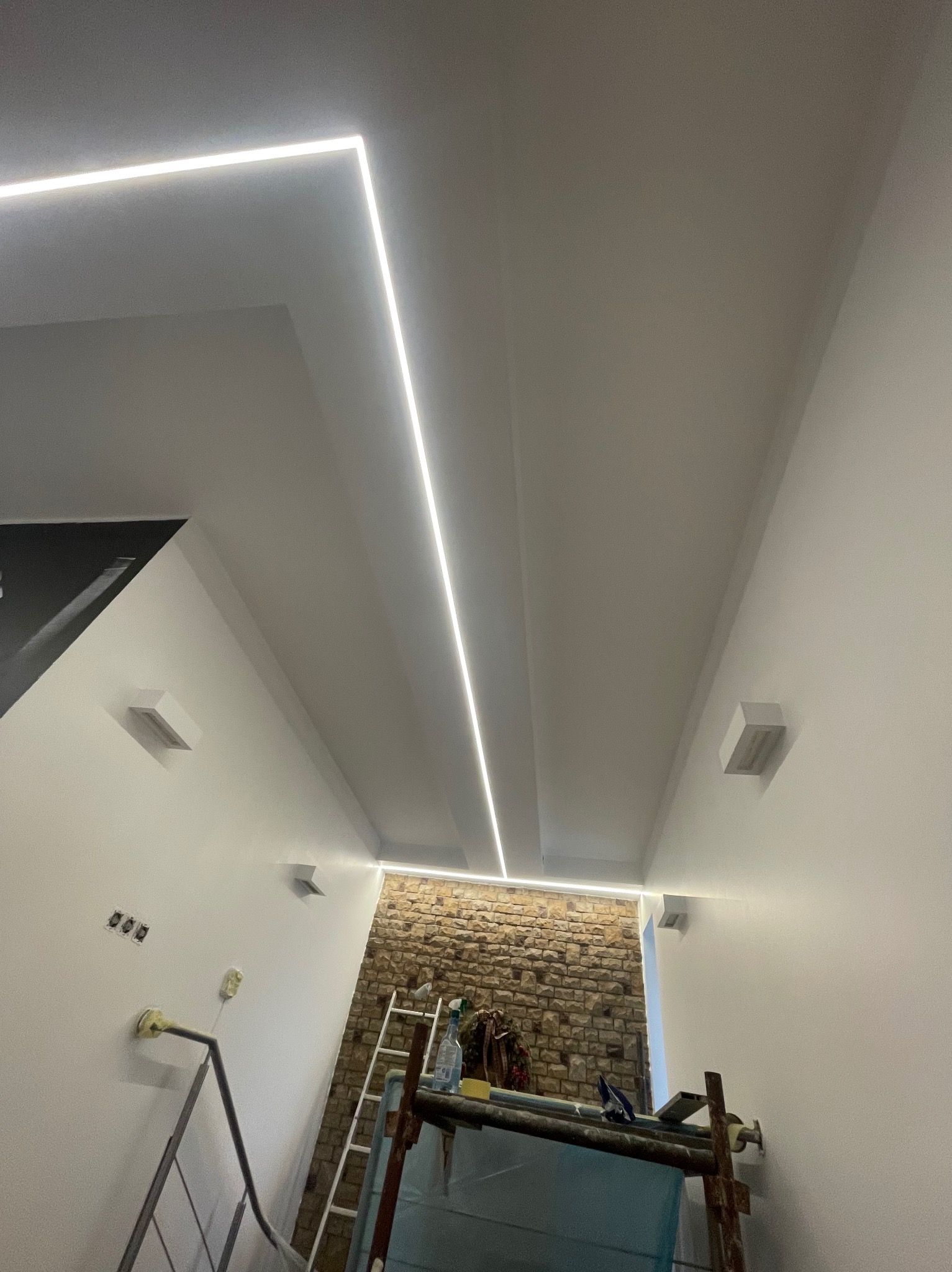Wnętrze w trakcie prac wykończeniowych: białe ściany, sufit z liniowym oświetleniem LED, fragment ściany z kamienia dekoracyjnego, widoczne rusztowanie i drabina.