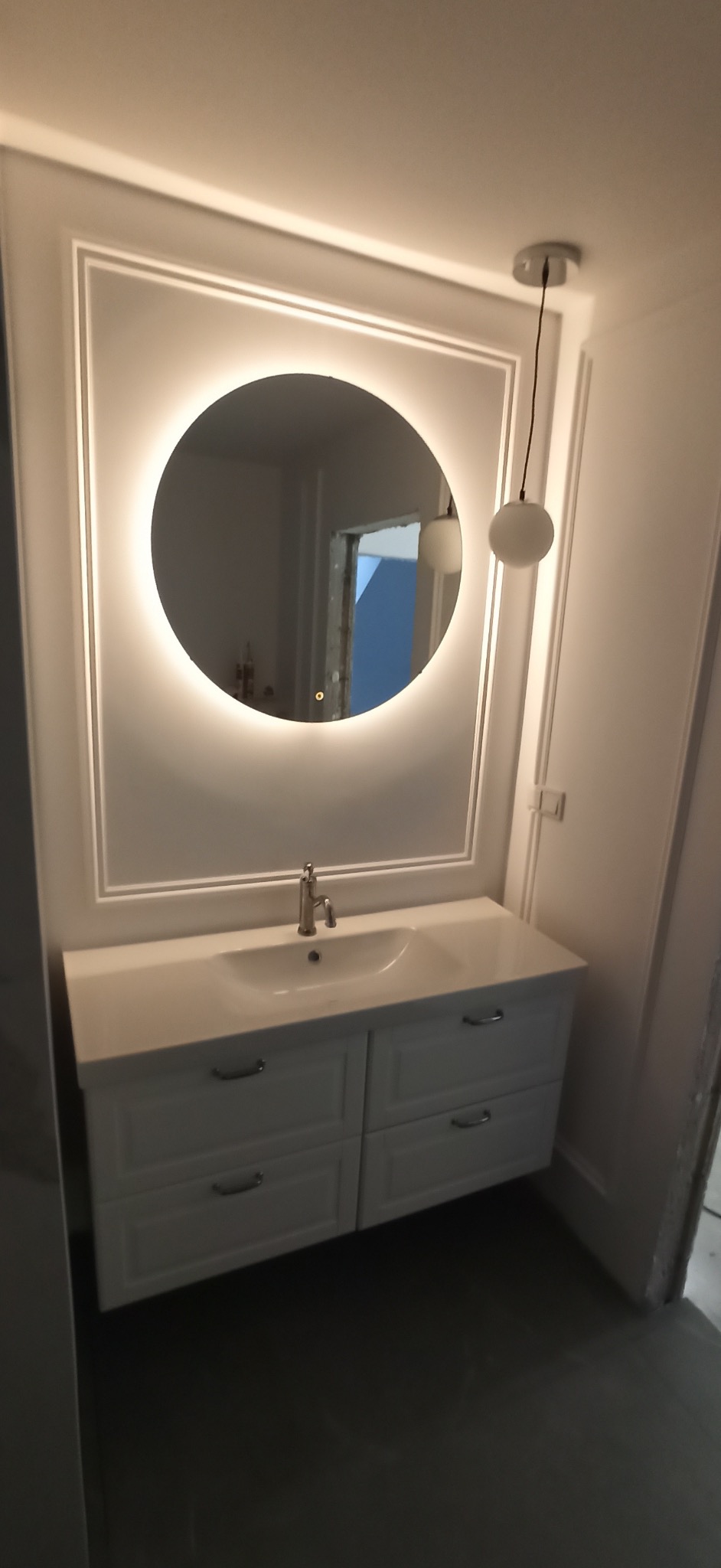 Biała umywalka z szafką podwieszaną, okrągłe lustro z podświetleniem LED w ozdobnej ramie, wisząca lampa kula, wykończenie ścian w stylu klasycznym.