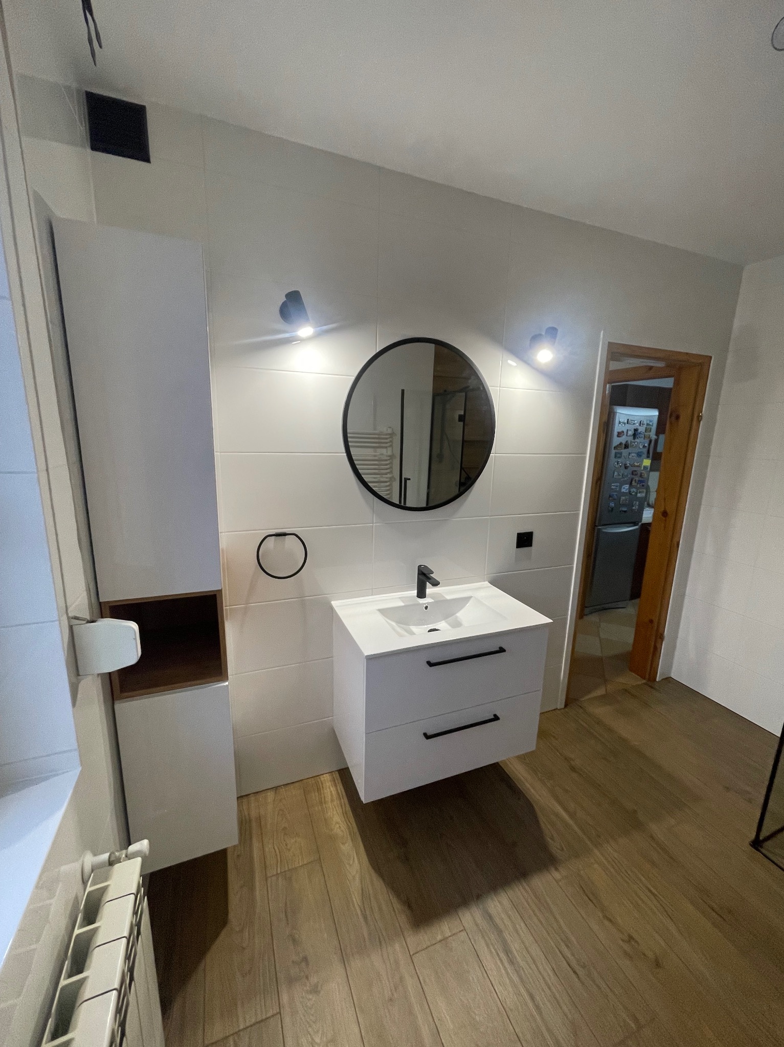 Nowoczesna łazienka w Śremie po liftingu, z białymi płytkami, drewnopodobną podłogą, okrągłym lustrem i minimalistyczną szafką z umywalką.