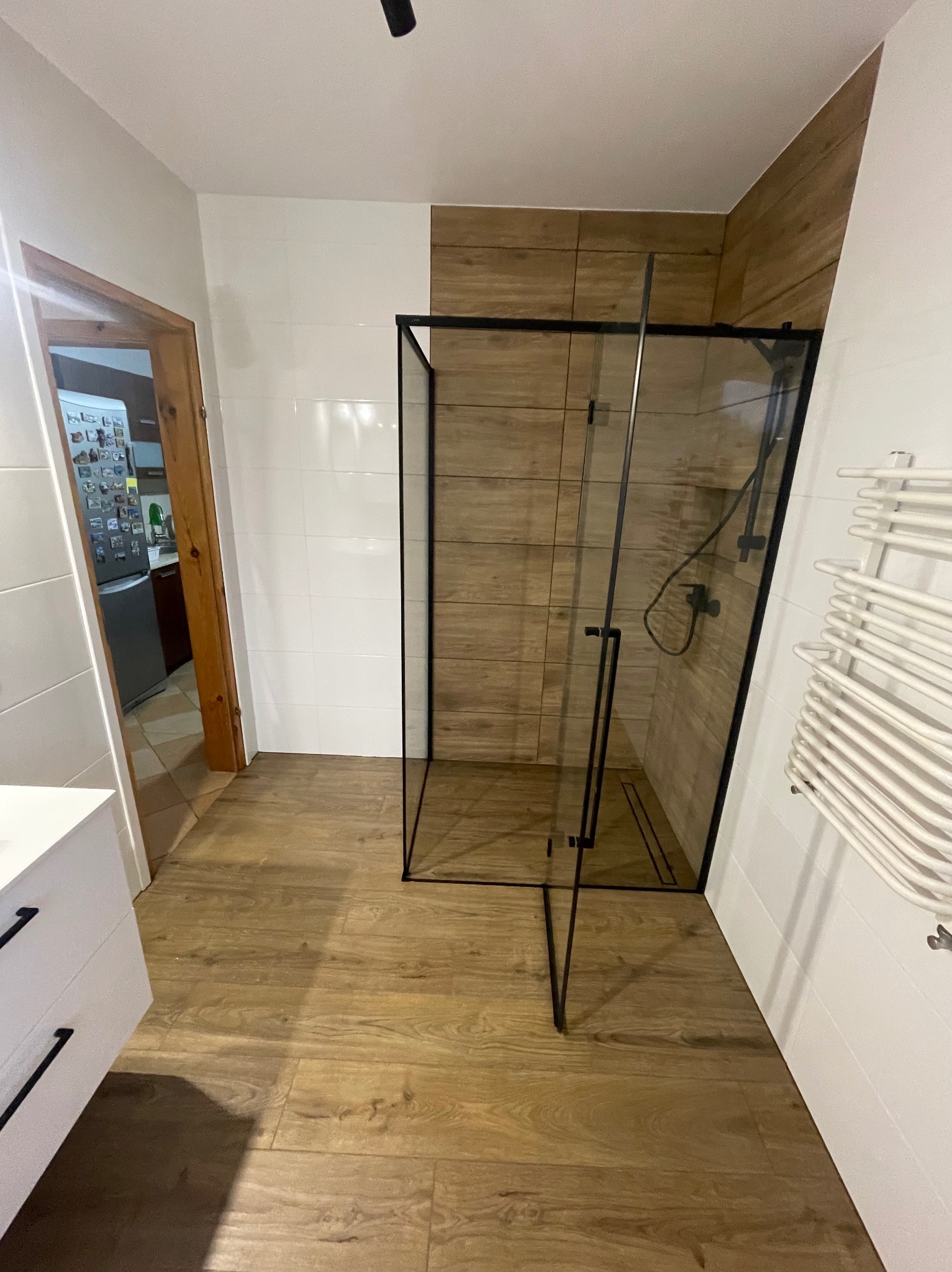 Nowoczesna łazienka z kabiną prysznicową typu walk-in z czarnymi profilami, drewnopodobnymi płytkami na ścianie i podłodze, oraz białym grzejnikiem drabinkowym.