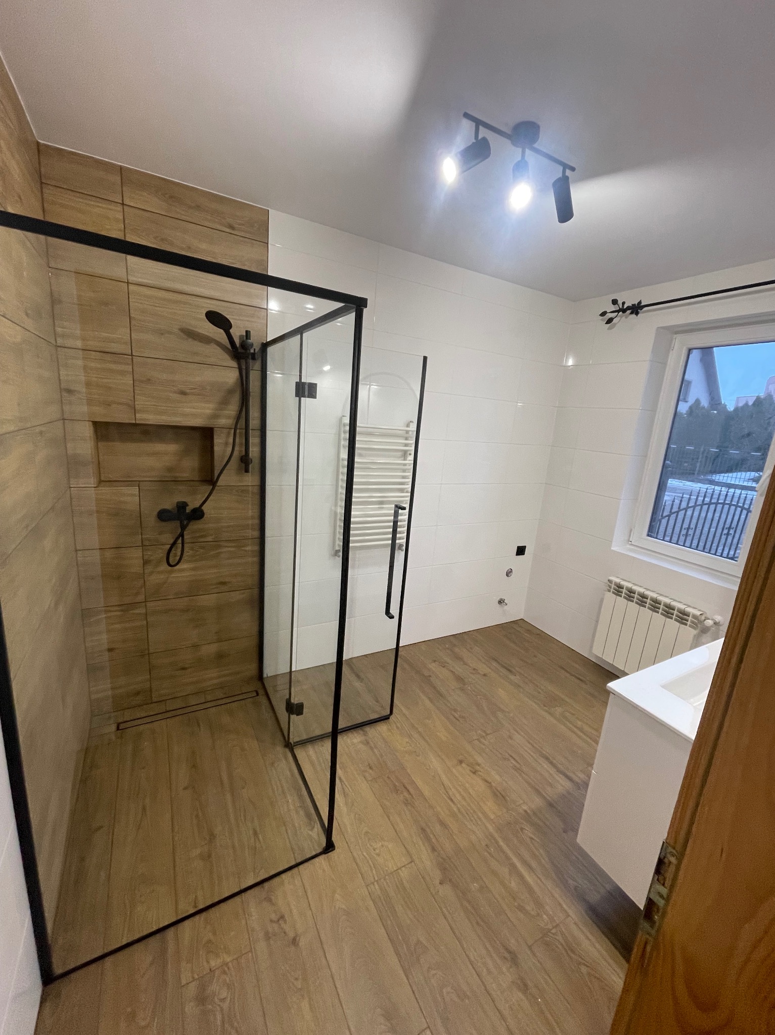 Nowoczesna łazienka z kabiną prysznicową typu walk-in z czarnymi profilami, drewnopodobne płytki w strefie prysznica i na podłodze, białe płytki na pozostałych ścianach, grzejnik drabinkowy, biała...