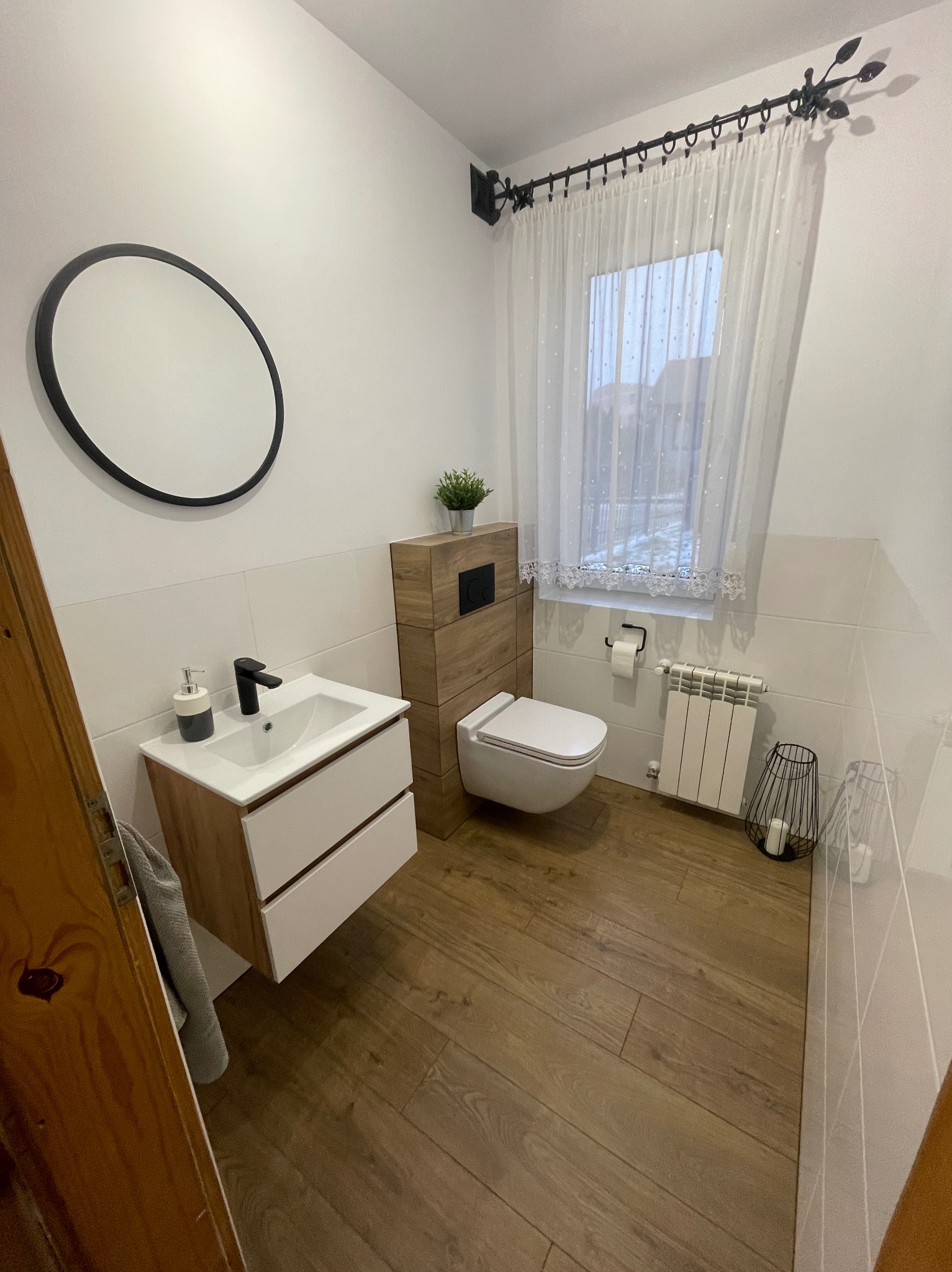 Nowoczesna łazienka z drewnianą podłogą i szafką pod umywalką, okrągłe lustro i zasłona w oknie.