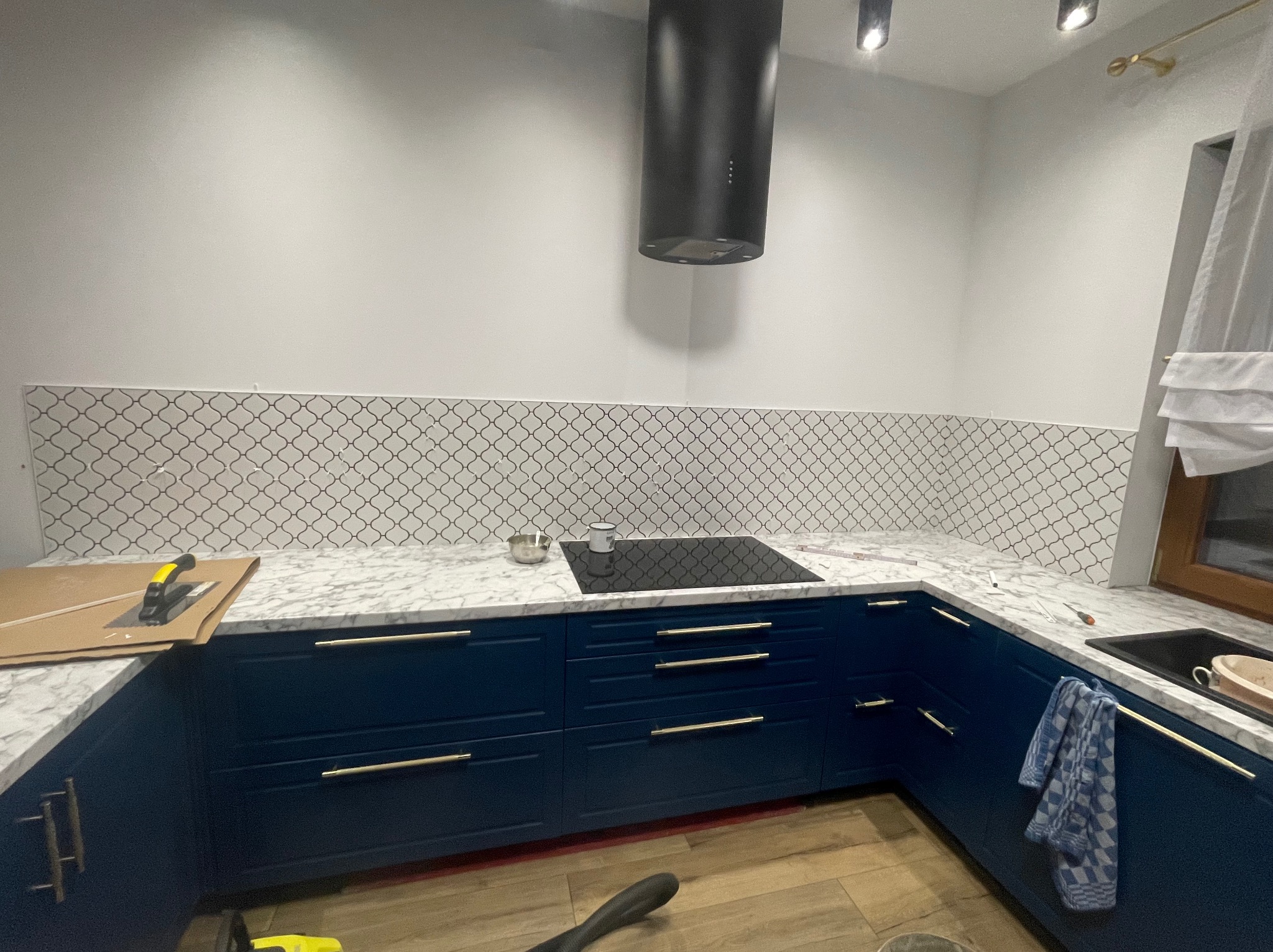 Niebieskie szafki kuchenne z marmurowym blatem i płytkami mozaikowymi na ścianie, czarny okap, narzędzia glazurnicze na blacie, widoczne etapy prac wykończeniowych.
