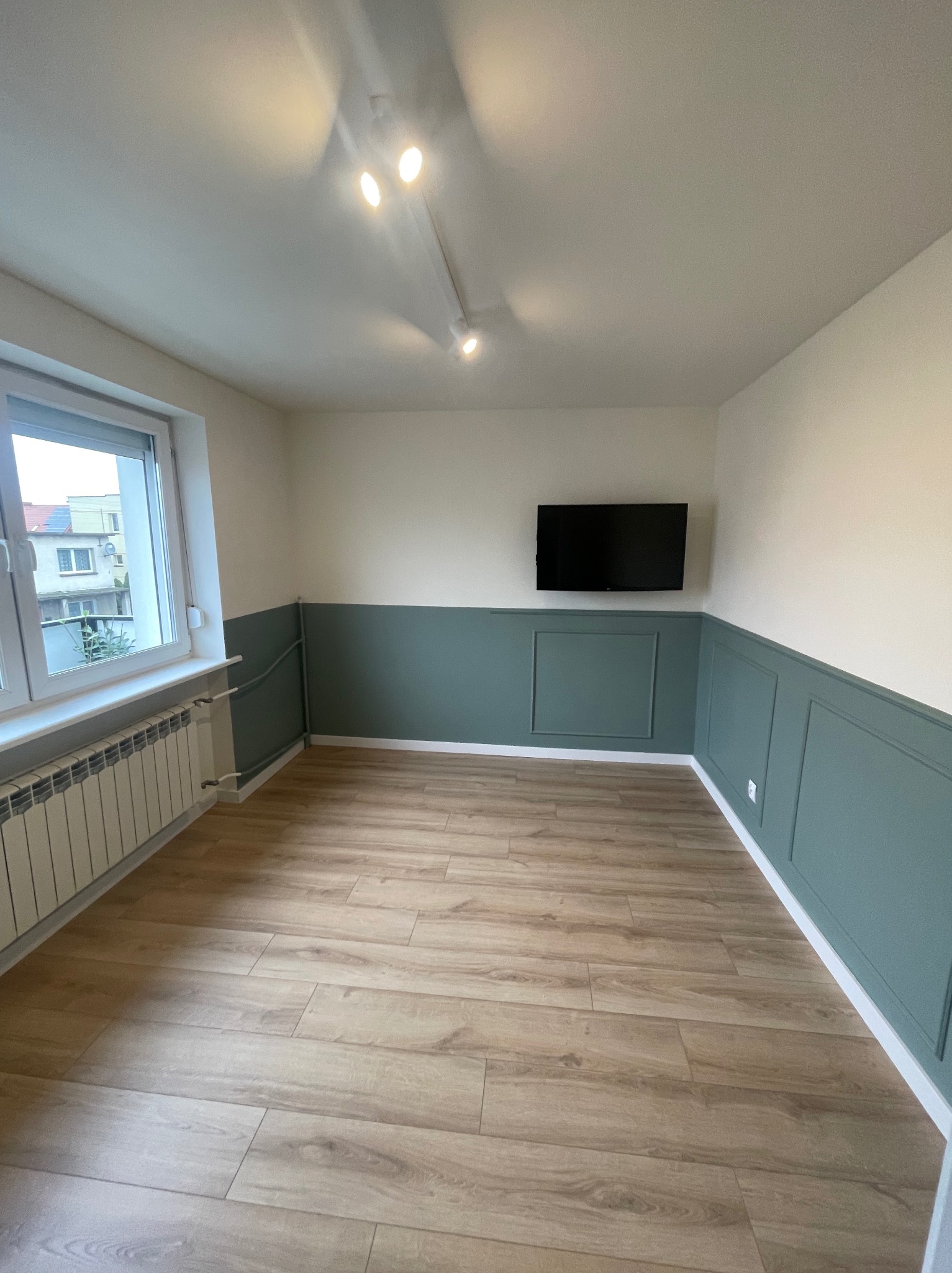 Jasne panele podłogowe w pokoju z pomalowanymi na zielono ścianami do połowy wysokości i zamontowanym telewizorem na ścianie.