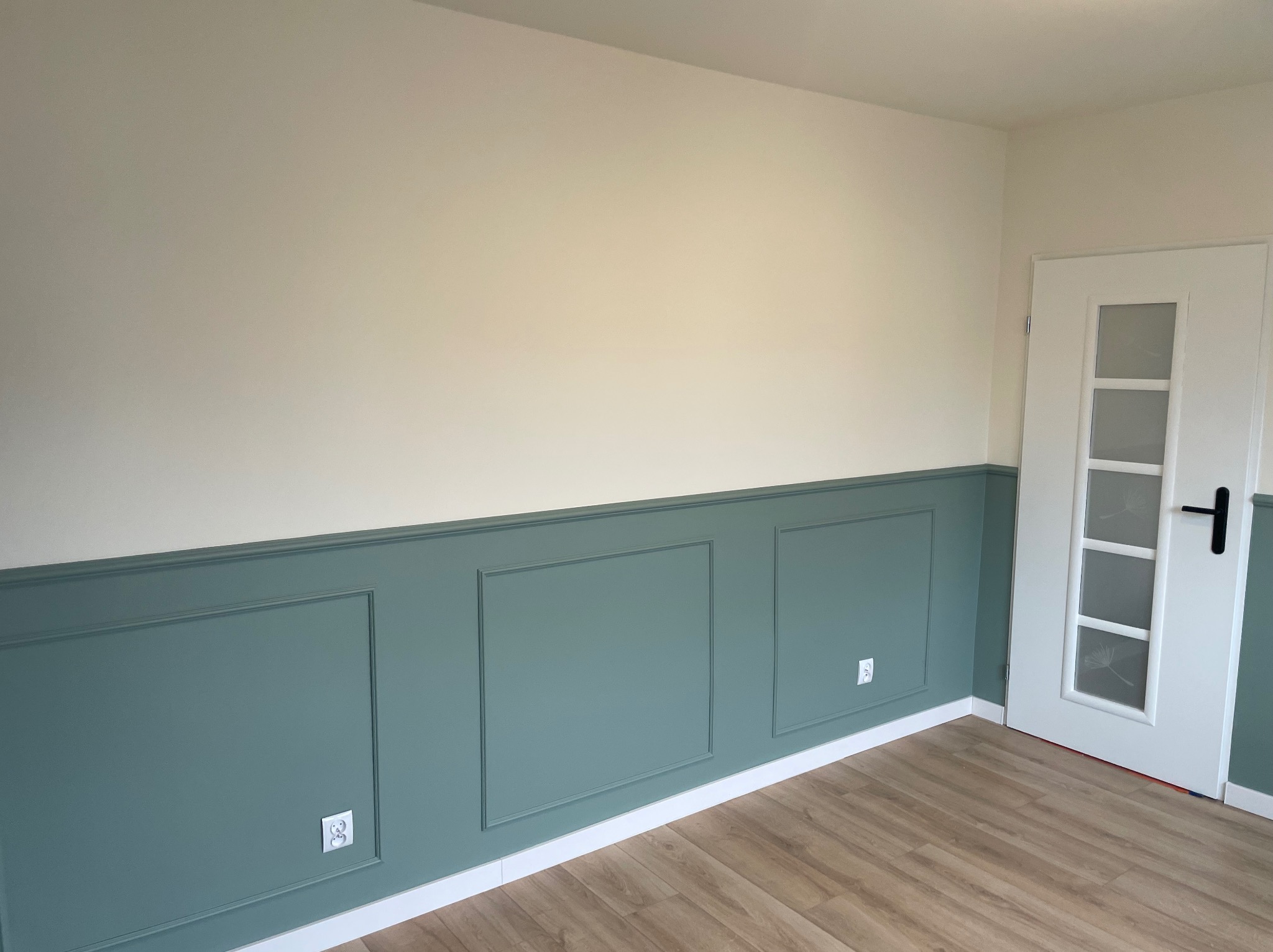 Wykończone wnętrze pokoju z panelami ściennymi w kolorze butelkowej zieleni i jasnymi ścianami, drewniana podłoga i białe drzwi z szybkami.