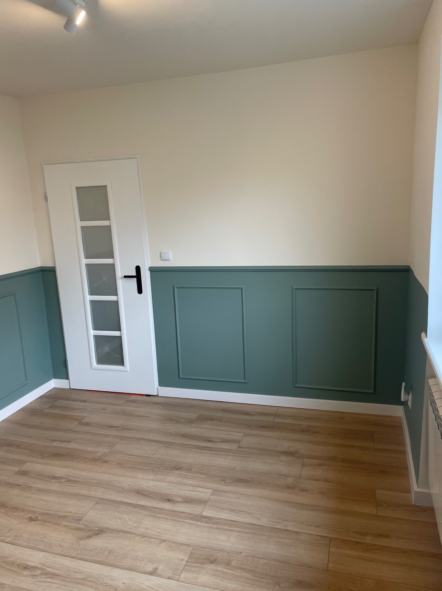 Wnętrze pokoju po remoncie: zielona lamperia na ścianach, drewnopodobna podłoga, białe drzwi z szybami z motywem dmuchawca.