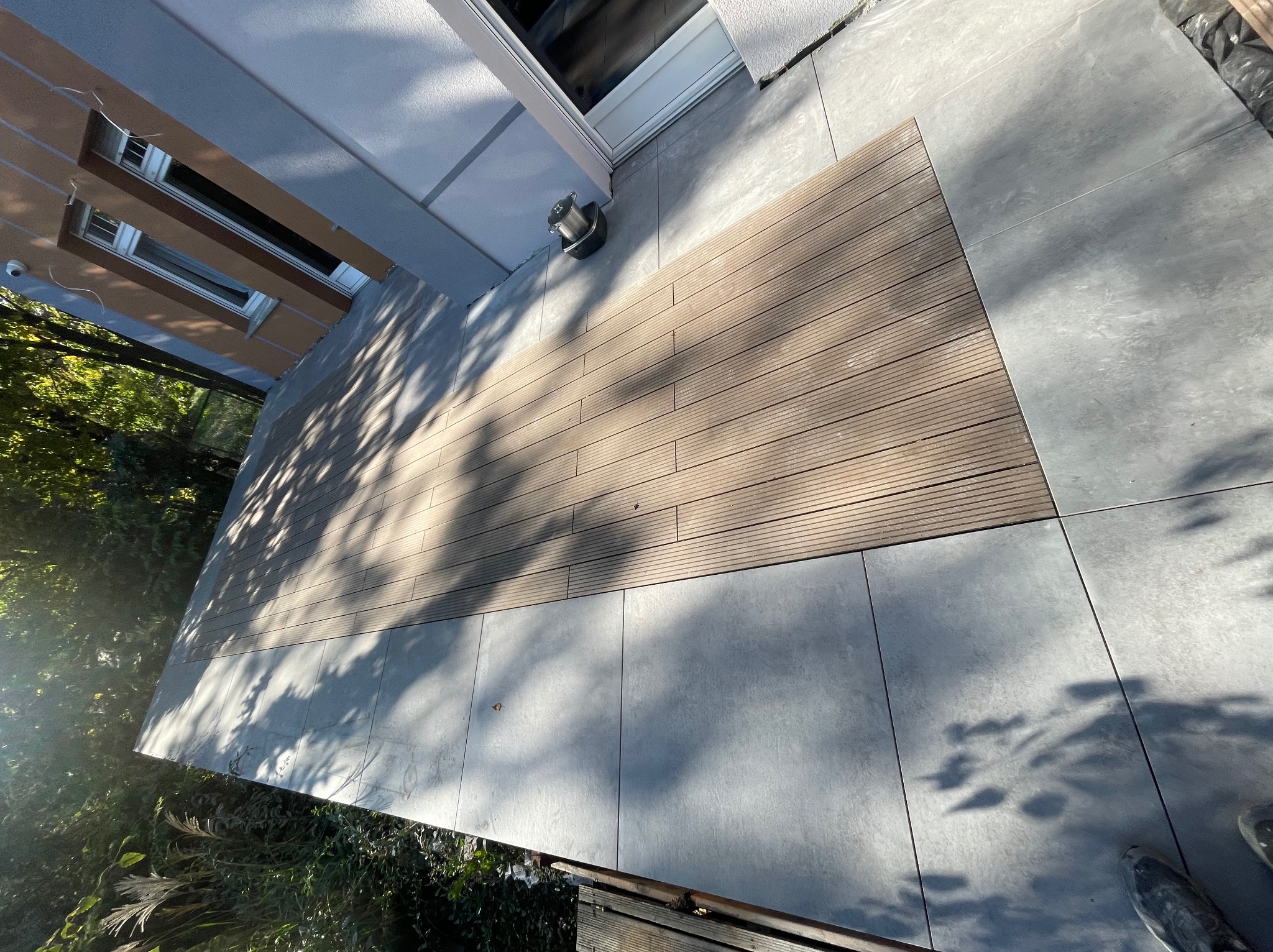 Wyłożony szarymi płytami i drewnianym tarasem fragment zewnętrzny domu z widocznymi cieniami drzew.