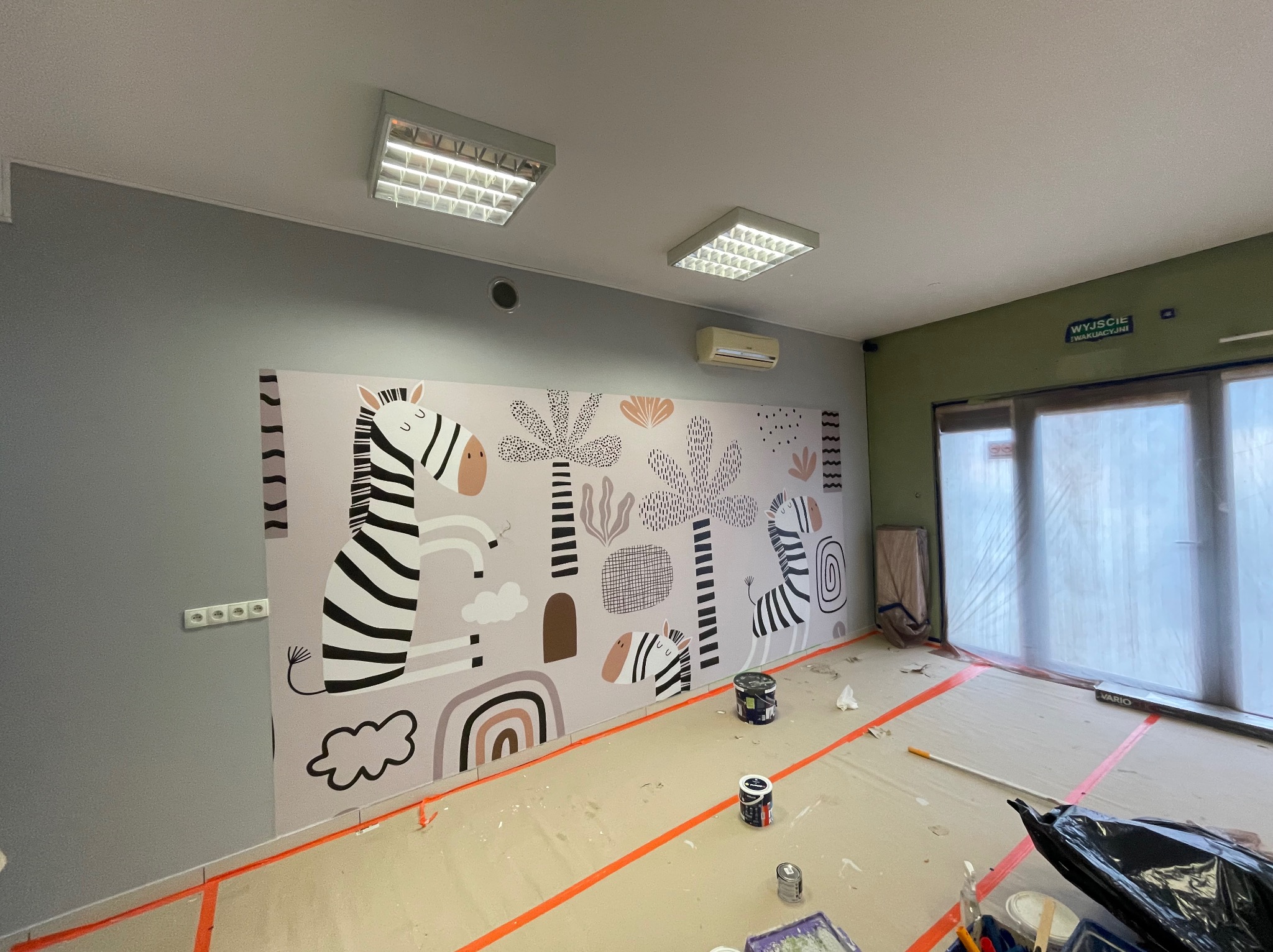 Pokój w trakcie malowania z motywem zebry i roślin na ścianie, widoczne puszki z farbą i taśma malarska na podłodze zabezpieczonej folią.