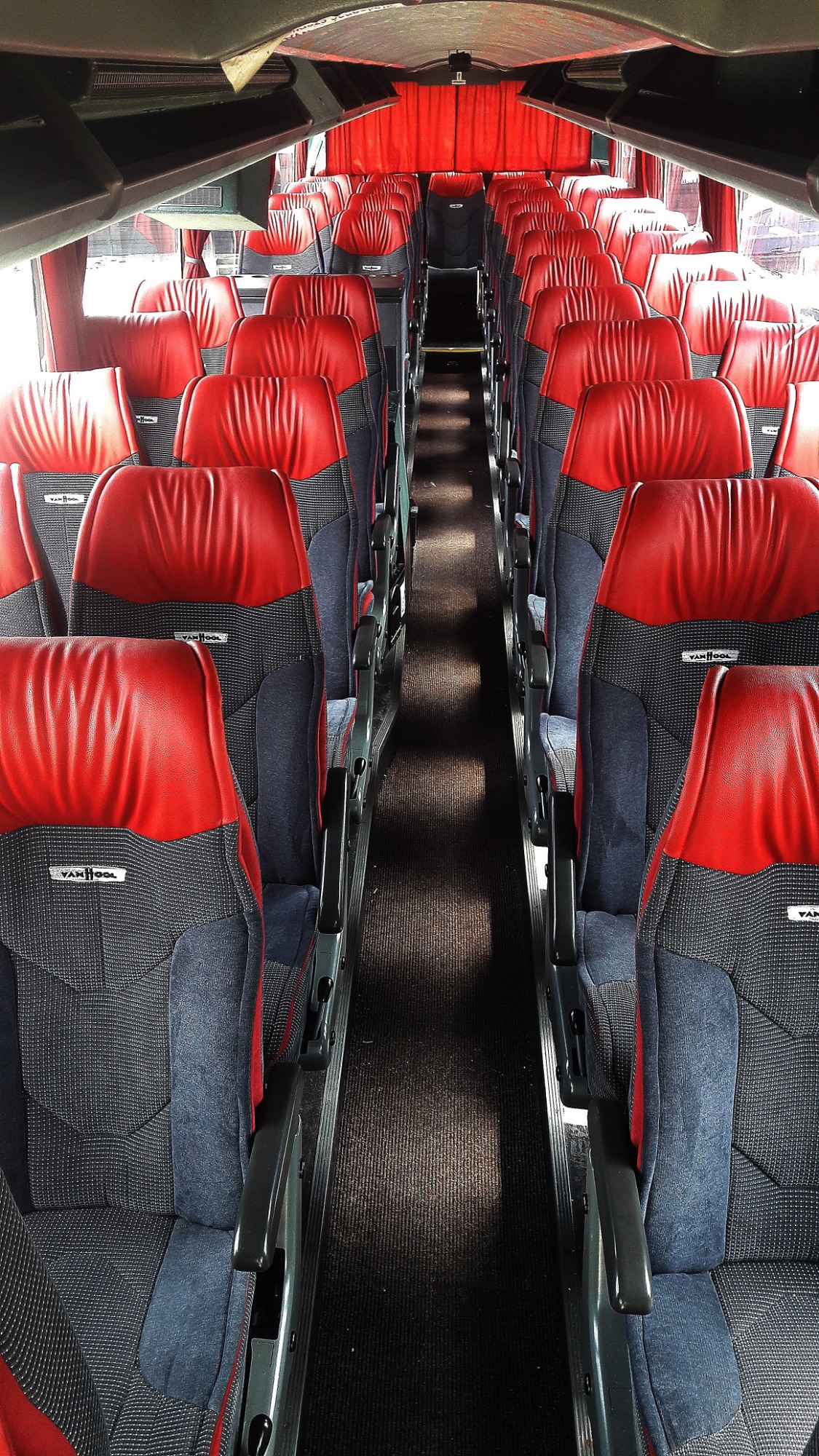 Wnętrze autobusu z rzędami czerwono-szarych foteli Van Hool, widok wzdłuż korytarza, czerwone zasłony na przedniej szybie.