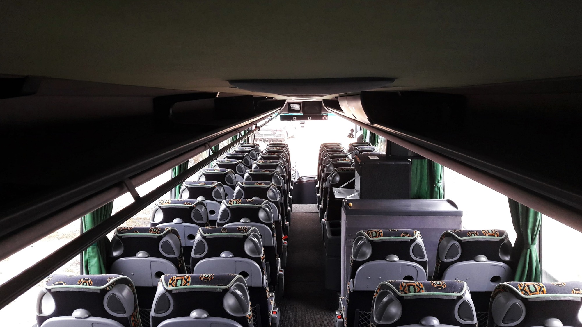 Wnętrze autobusu turystycznego, widok z tyłu na rzędy foteli z tapicerką w geometryczne wzory, zielone zasłony przy oknach.