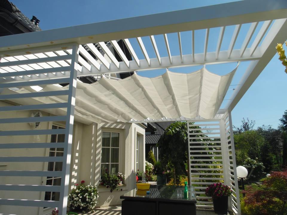 Biała pergola metalowa z rozsuwanym, falistym zadaszeniem materiałowym, widok z niskiej perspektywy na tle błękitnego nieba i częściowo zarośniętego ogrodu.