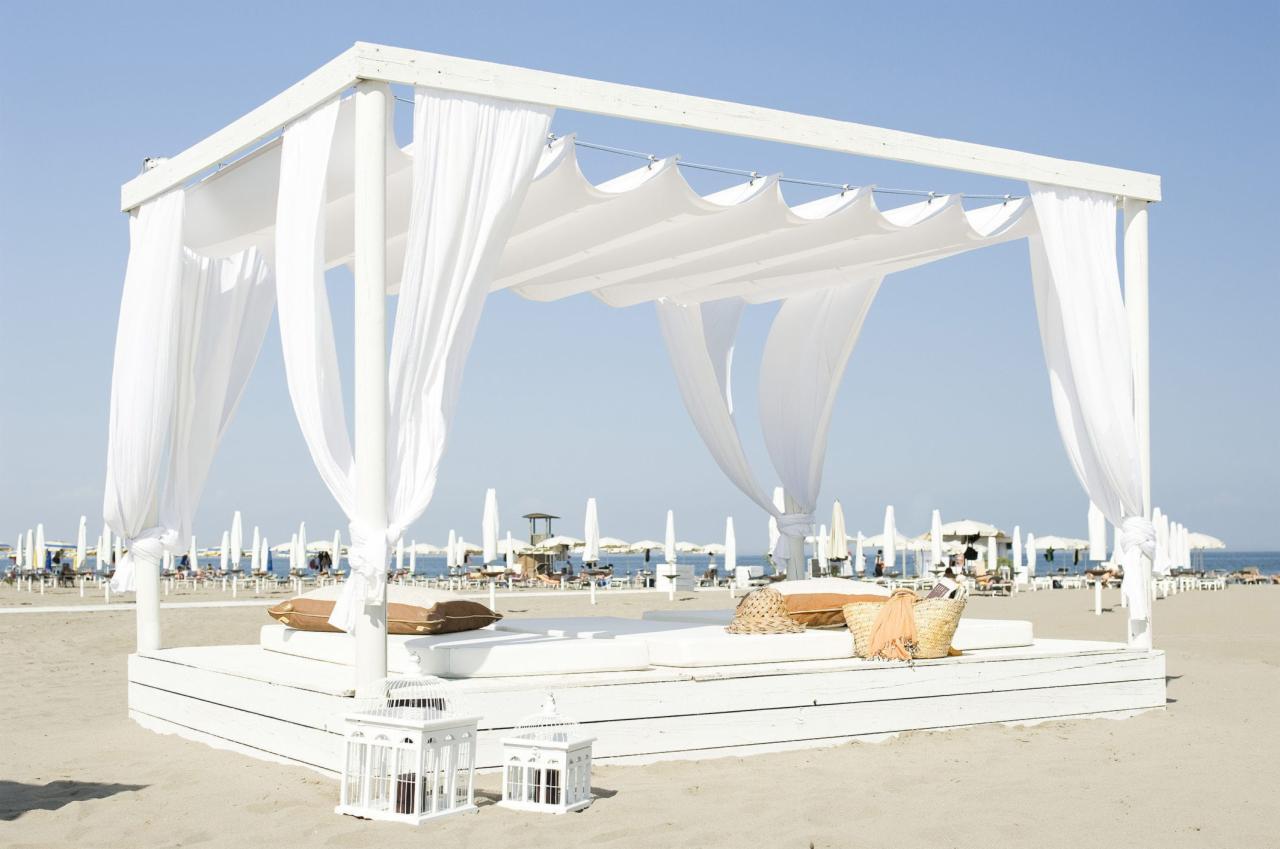 Biała, drewniana pergola z baldachimem na plaży, z widokiem na morze i parasole w tle, dekorowana poduszkami, koszykiem i lampionami.