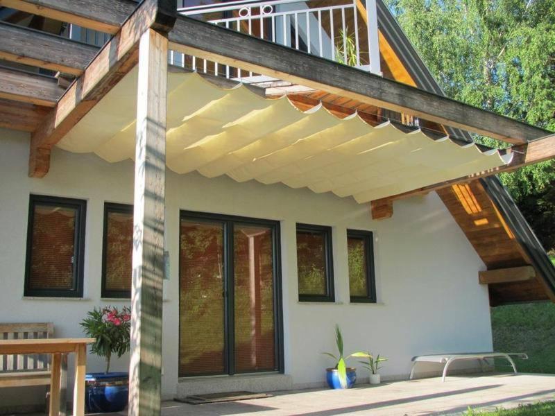 Drewniana konstrukcja z rozsuwanym, kremowym zadaszeniem nad tarasem przy białym domu z ciemnymi oknami i drzwiami przesuwnymi.