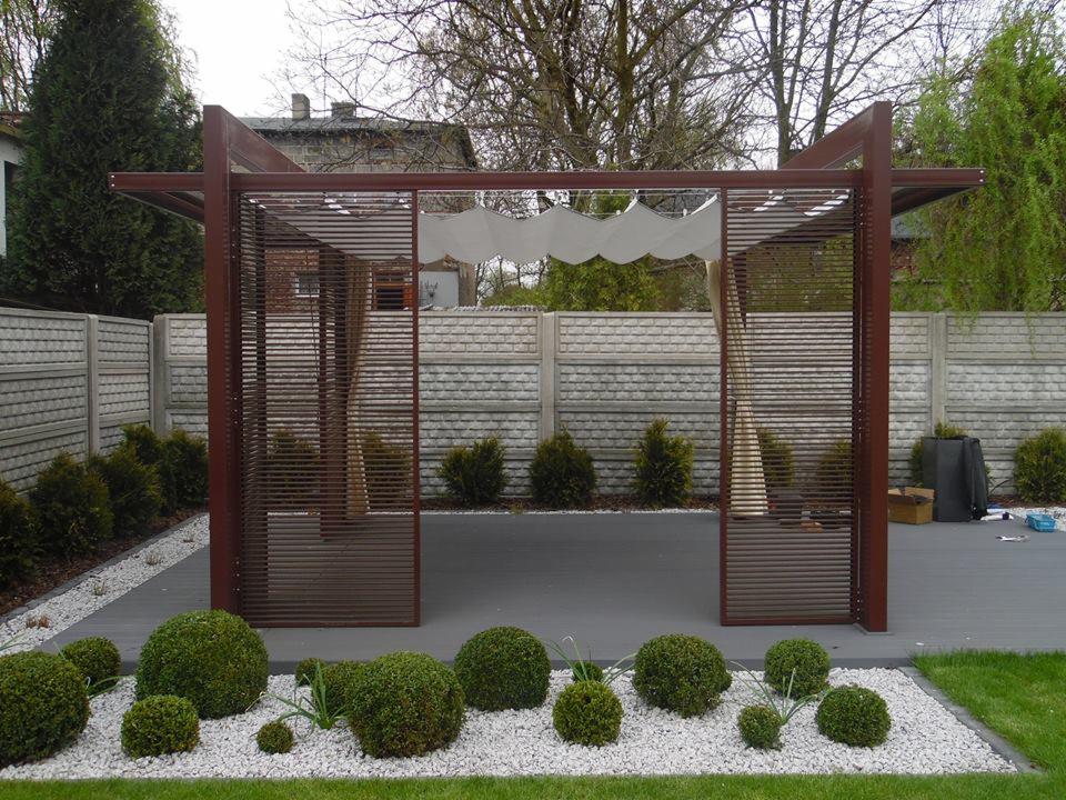 Nowoczesna pergola z brązowych profili z poziomymi listwami, dekoracyjna biała tkanina w górnej części, otoczona kulistymi krzewami na białym żwirze, szary taras i betonowe ogrodzenie w tle.