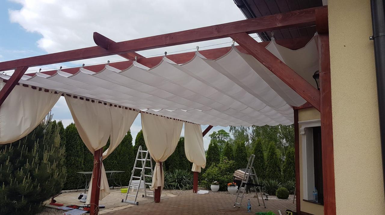 Drewniana konstrukcja z beżowymi zasłonami na systemie szynowym, dwie drabiny opierają się o konstrukcję, w tle iglaki i kostka brukowa.