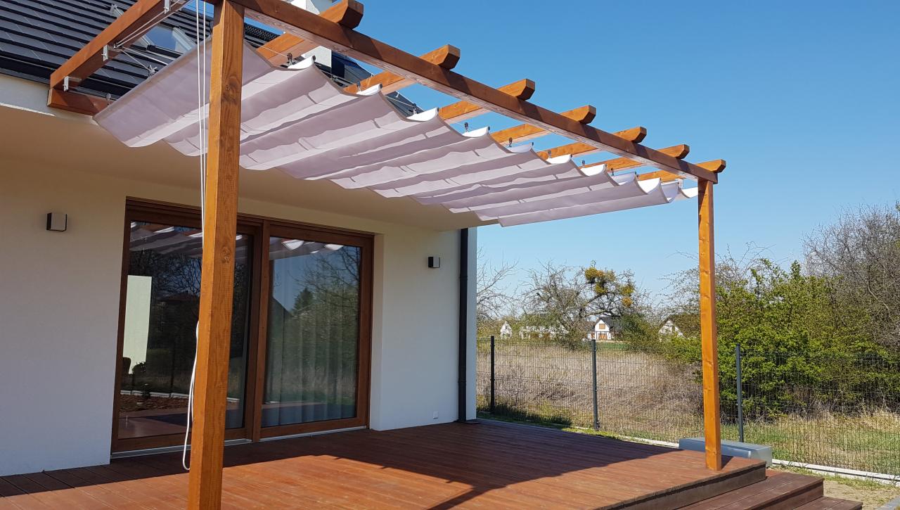 Drewniana pergola z rozsuwanym, jasnym zadaszeniem materiałowym, stojąca na tarasie z desek, przylegająca do białego domu z dużymi, przesuwnymi oknami.