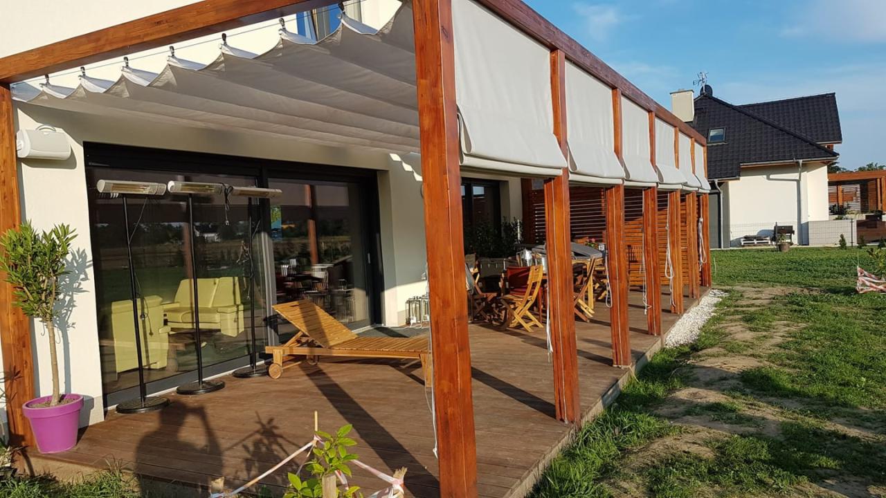 Drewniana pergola z rozsuwanym, kremowym zadaszeniem nad tarasem z desek. Widoczne drewniane meble ogrodowe, leżak i trzy grzejniki tarasowe. W tle dom z ogrodem.