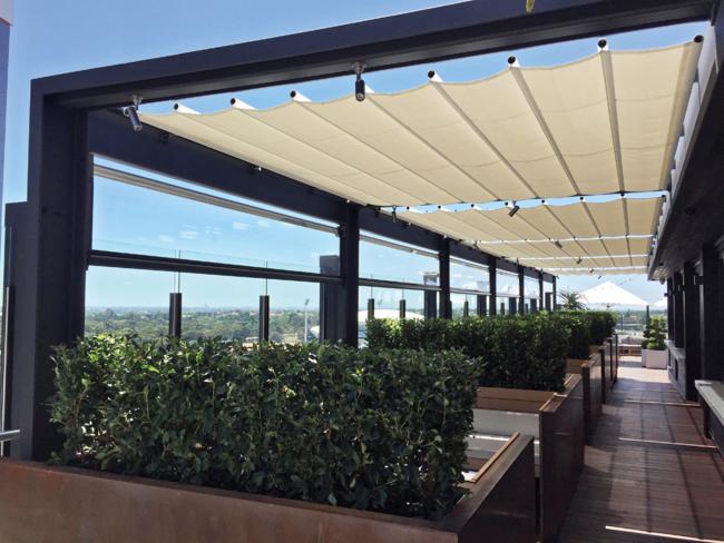 Zadaszony taras na dachu w Gościęcicach z pergolą z metalową konstrukcją i rozsuwanym beżowym płótnem, osłonięty szklaną balustradą i zielenią w donicach.