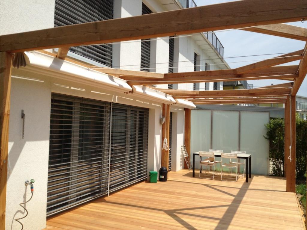 Drewniany taras z pergolą z surowego drewna i markizą przy budynku z żaluzjami, stół z krzesłami na tarasie, zielony krzew w tle.