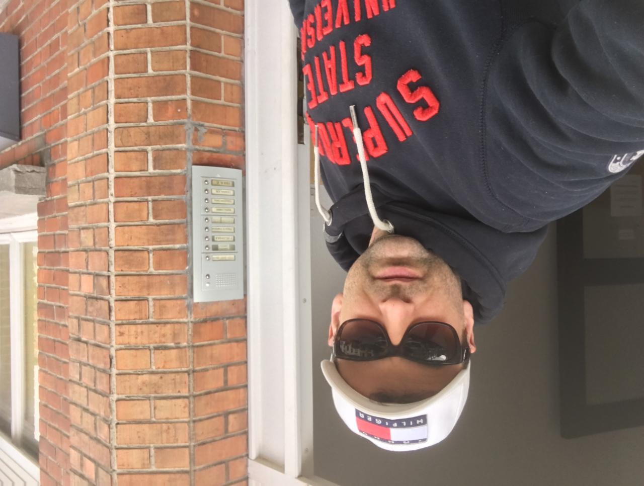 Mężczyzna w okularach i czapce Tommy Hilfiger stoi przed budynkiem z cegły z domofonem.