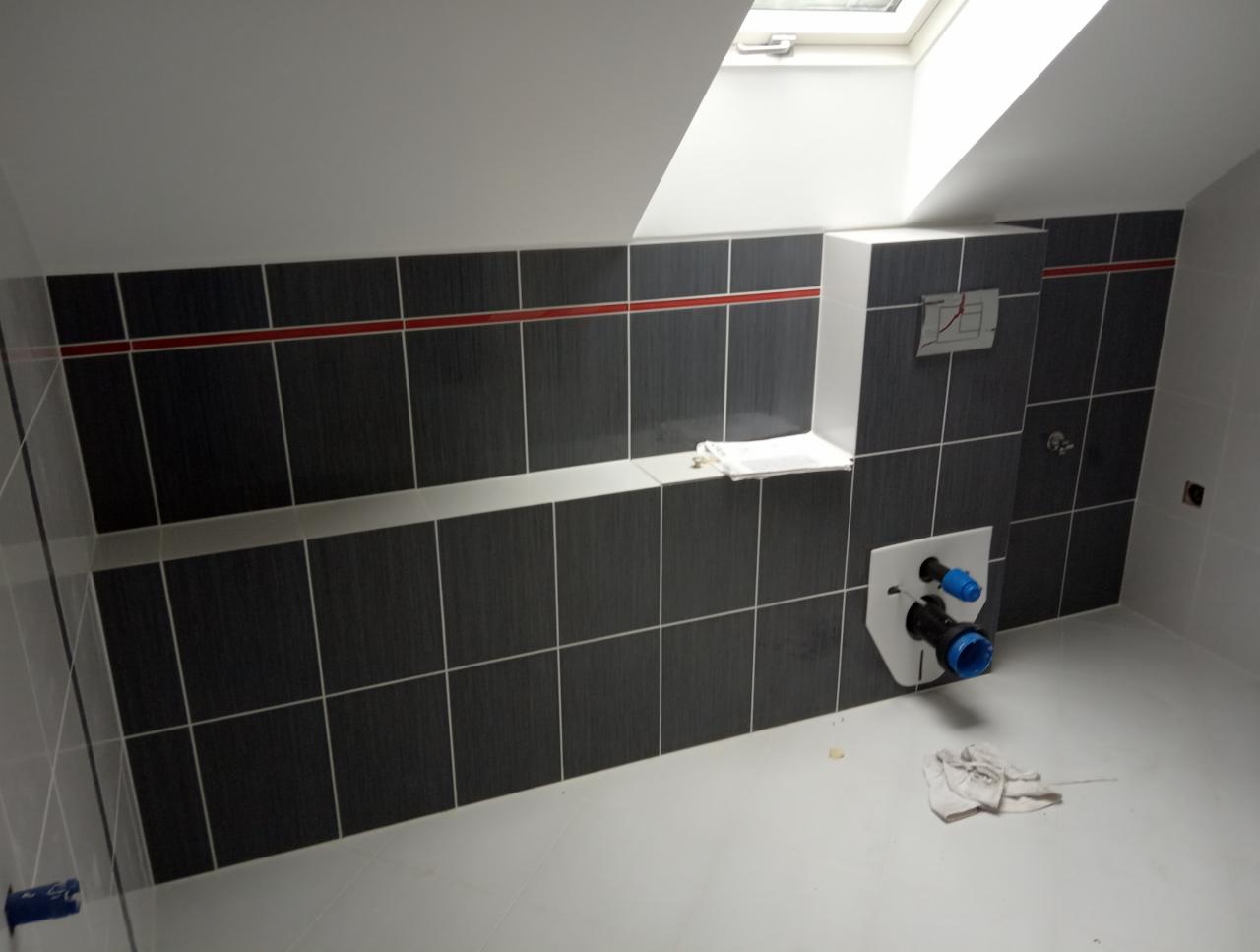 Łazienka w trakcie remontu na poddaszu: ciemne płytki z czerwoną linią, biała podłoga i stelaż podtynkowy WC z niebieskimi elementami, widoczne okno dachowe.