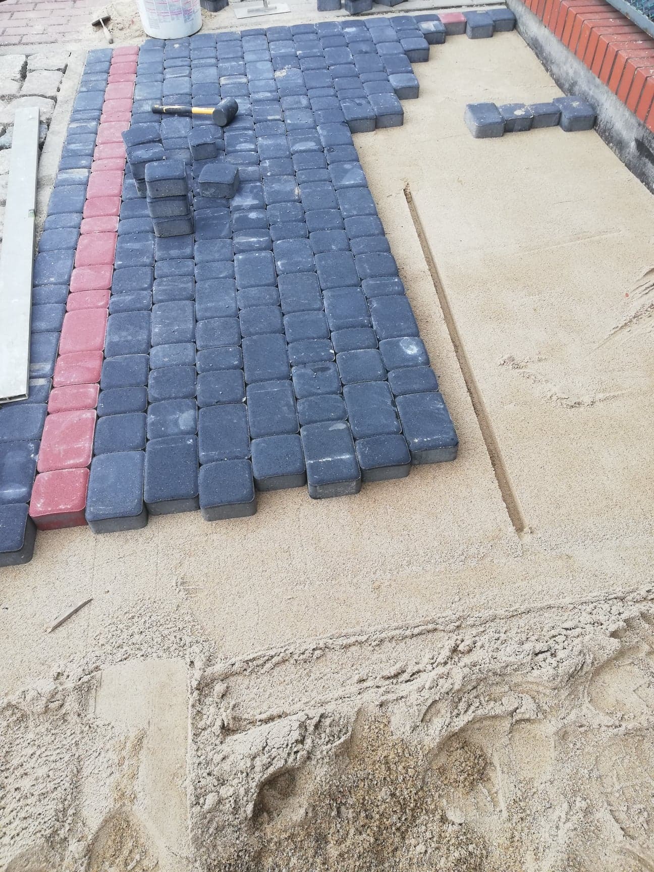 Fragment układanej nawierzchni z ciemnoszarej kostki brukowej z czerwoną linią brzegową, widoczny gumowy młotek i stos kostek na piaskowym podłożu, etap prac.
