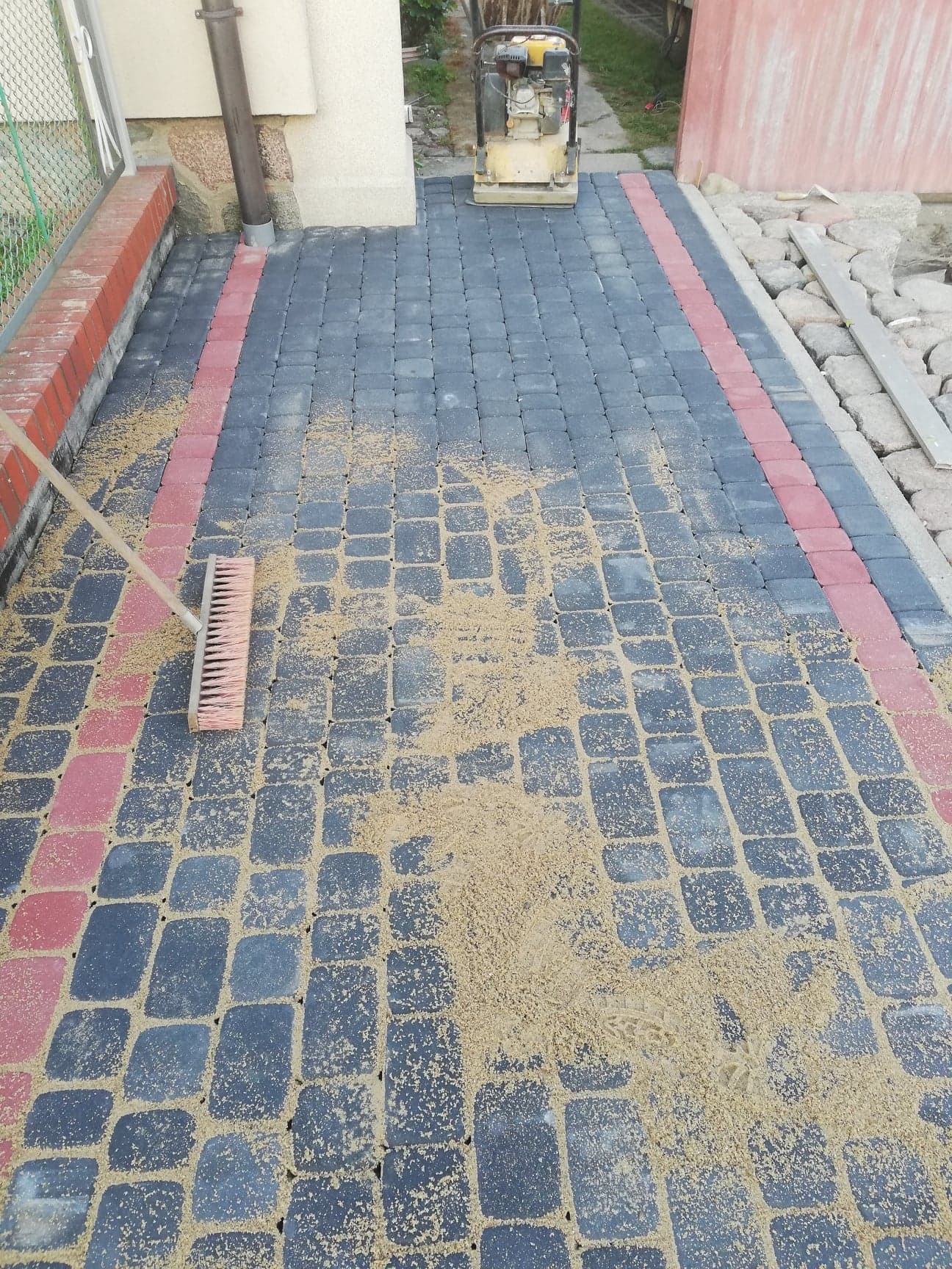 Szczotka zamiata piasek na nowo ułożonej nawierzchni z ciemnoszarej kostki brukowej z obramowaniem z czerwonej kostki; w tle ubijarka wibracyjna.
