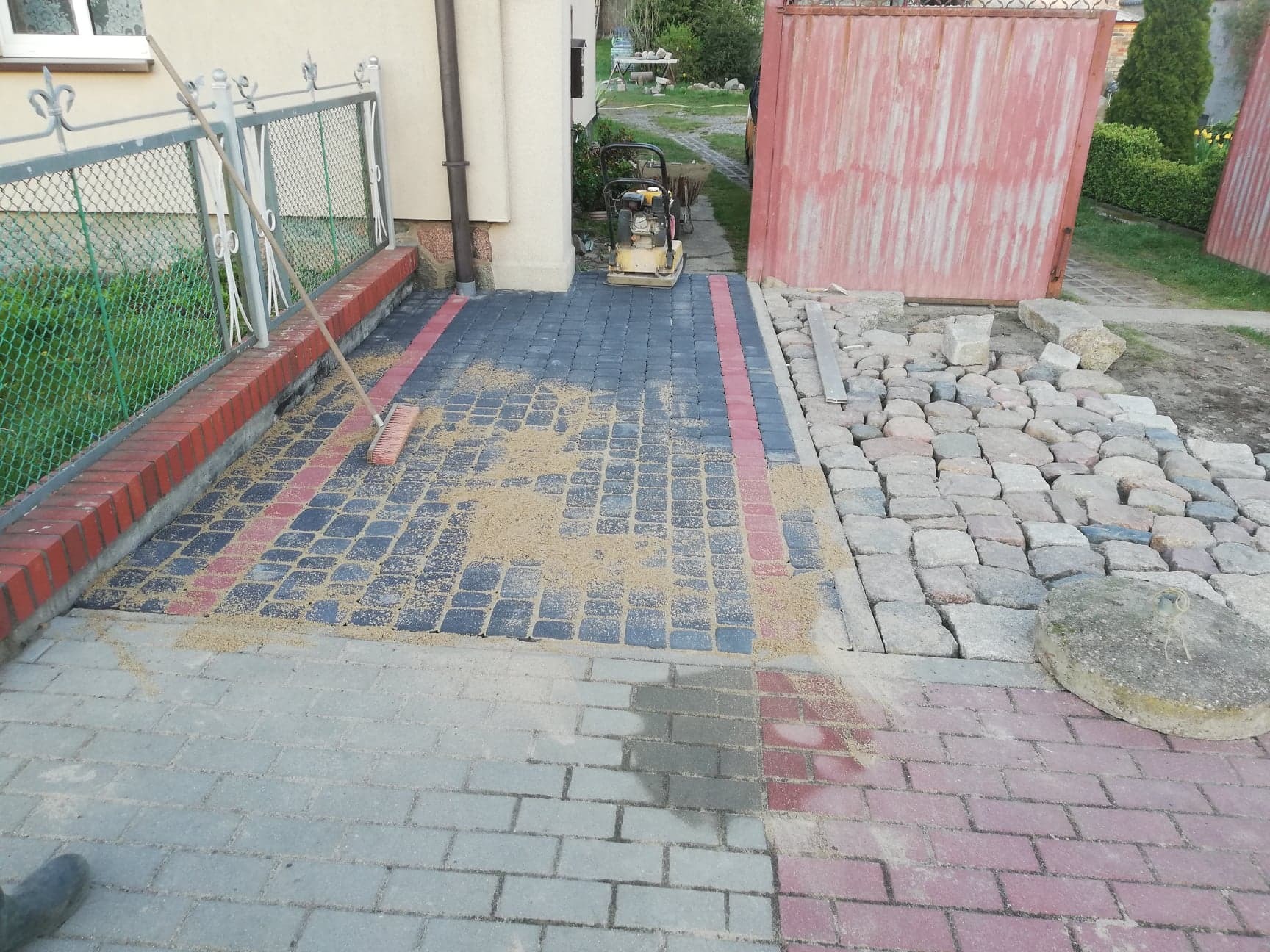 Fragment podwórza z częściowo ułożoną kostką brukową w trzech kolorach, piaskiem do spoinowania, ubijarką wibracyjną i przygotowanym fragmentem z kostki granitowej.